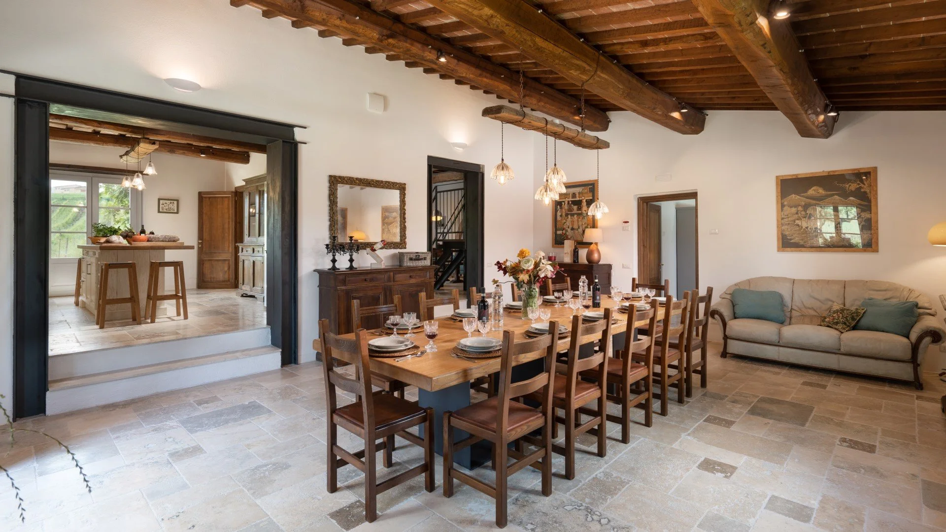 Dining room with a long wooden table set with dishes and glasses, surrounded by wooden chairs, beige tiled floor, white walls, wooden ceiling beams, and a beige sofa with pillows in the background.