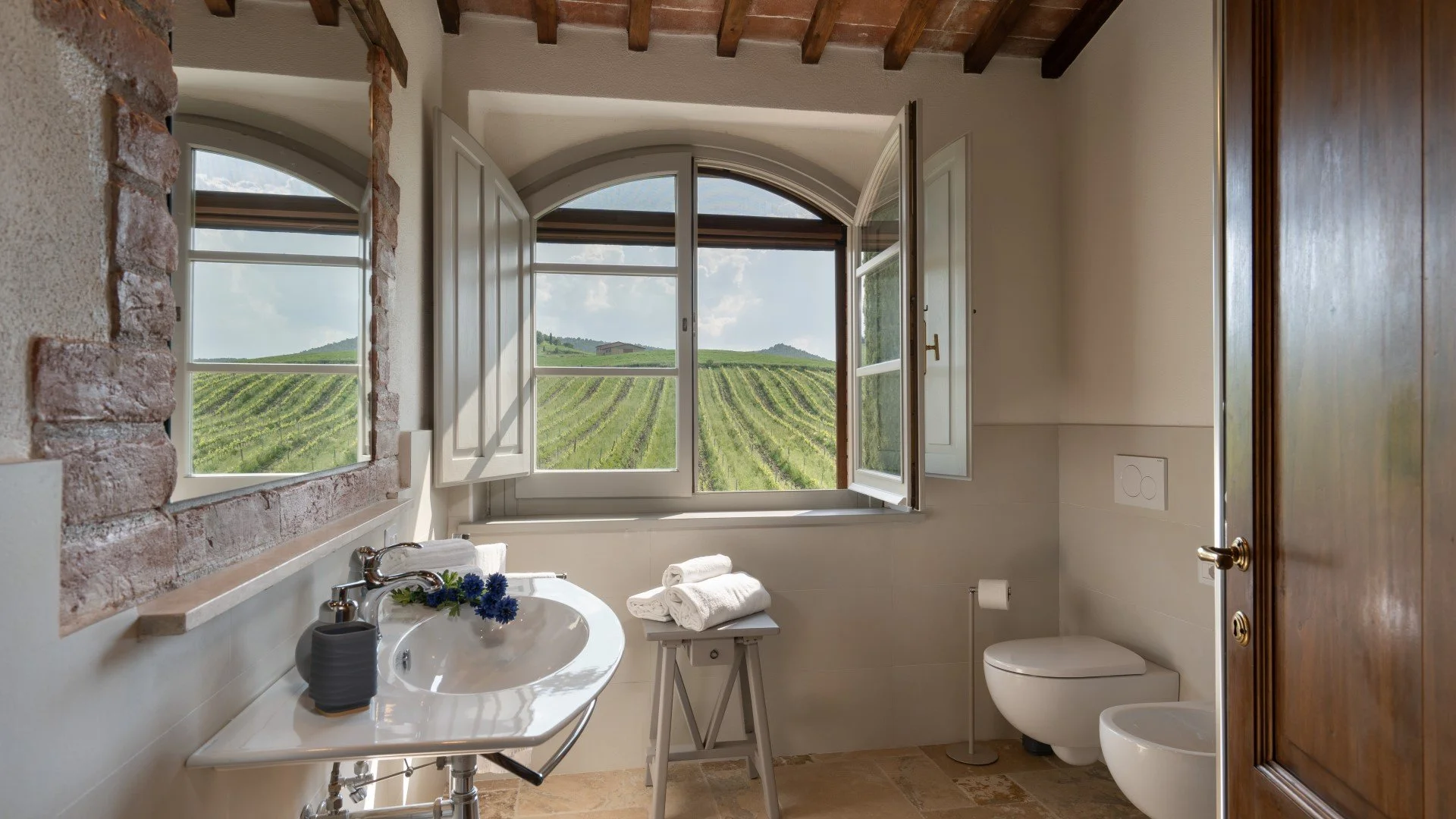 Bathroom with open window overlooking green fields and rolling hills, featuring a sink with a flower arrangement, folded towels, and a toilet.