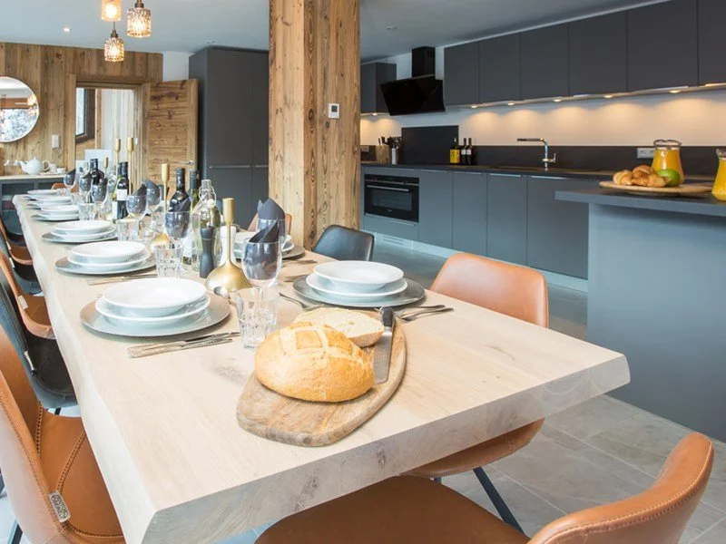 Modern dining room with a long wooden table set with white plates, glasses, and bottles, and a bread loaf on a wooden cutting board, in an open kitchen with gray cabinets and black countertops.