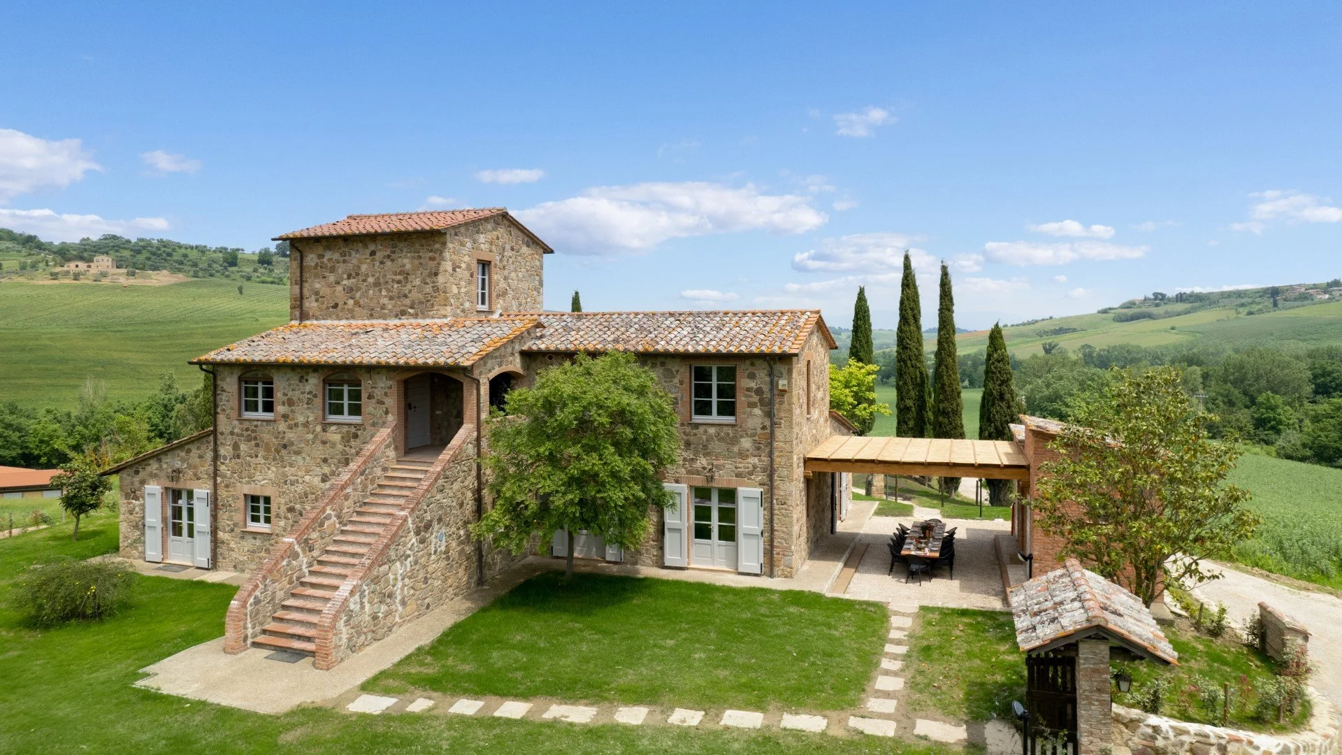 A rustic stone house with tiled roof, surrounded by green lawns and trees, and overlooking rolling hills under a blue sky with clouds.