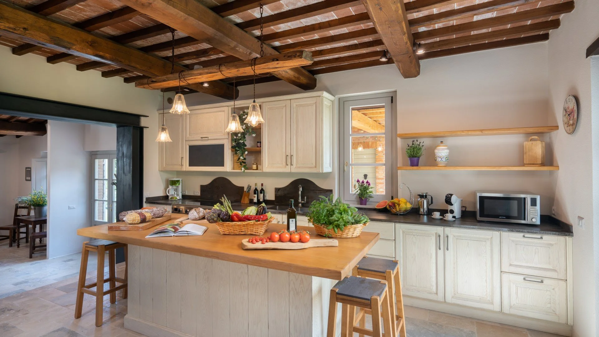 Kitchen with a wooden island, fresh vegetables, fruits, and a basket of tomatoes, cream-colored cabinets, open shelves with decorative items, and a window with natural light.