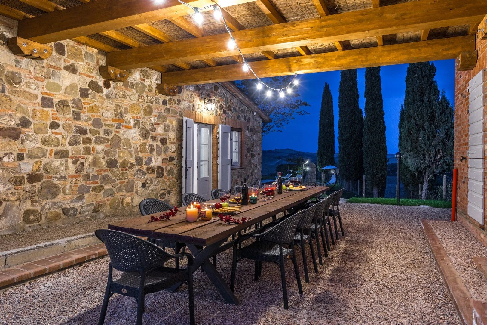 An outdoor dining area with a long wooden table set for a dinner, featuring candles, wine, and candles, under a wooden pergola with string lights, against a stone and brick wall, with tall cypress trees visible in the background at dusk.