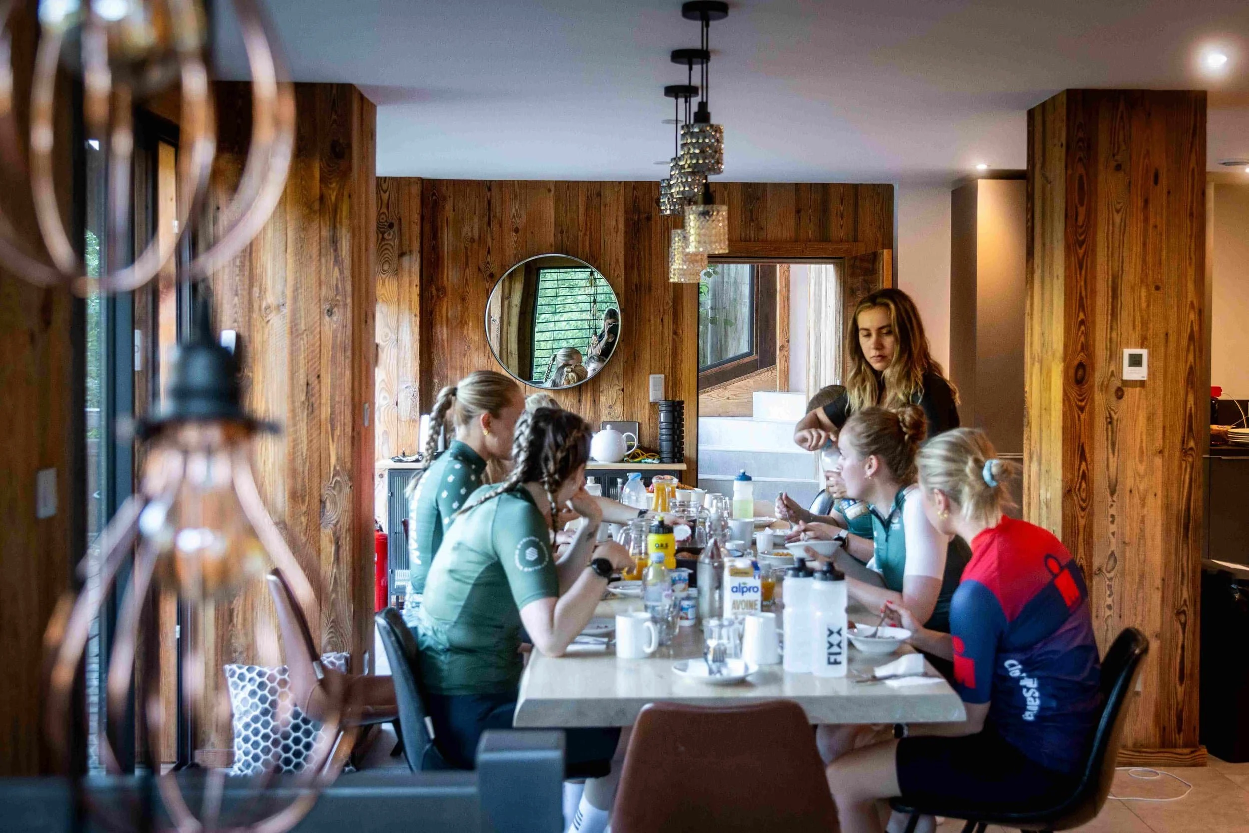 A group of women in cycling gear sitting at a wooden dining table in a cozy, rustic room with wood-paneled walls, having a meal or preparing for a gathering, with one woman standing and talking to the others.