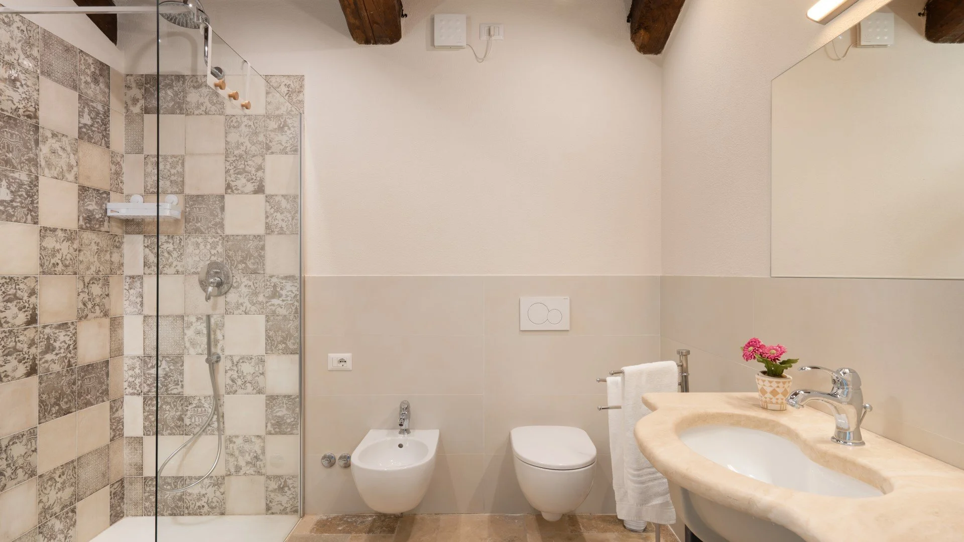 Bathroom with a walk-in shower featuring patterned tiles, a bidet, a toilet, a sink with a marble countertop, a mirror, a towel rack with a towel, and a small potted pink flower.