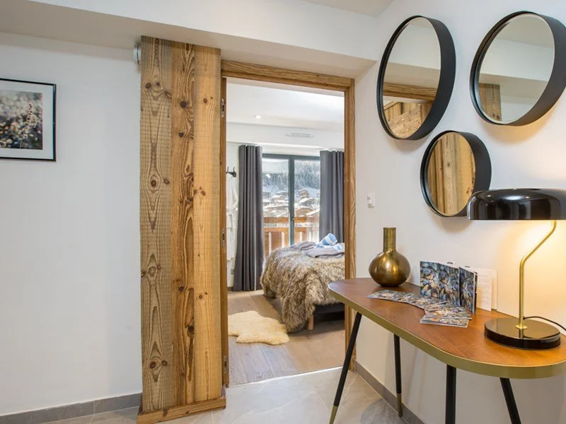 Interior view of a modern bedroom with large window, wooden sliding door, and cozy bedding, seen from an adjacent room with decorative mirrors and a side table.