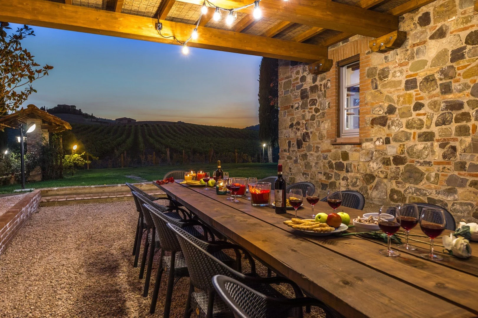 An outdoor dining table set for a meal at sunset, with wine glasses, bottles, and food, overlooking a vineyard landscape.
