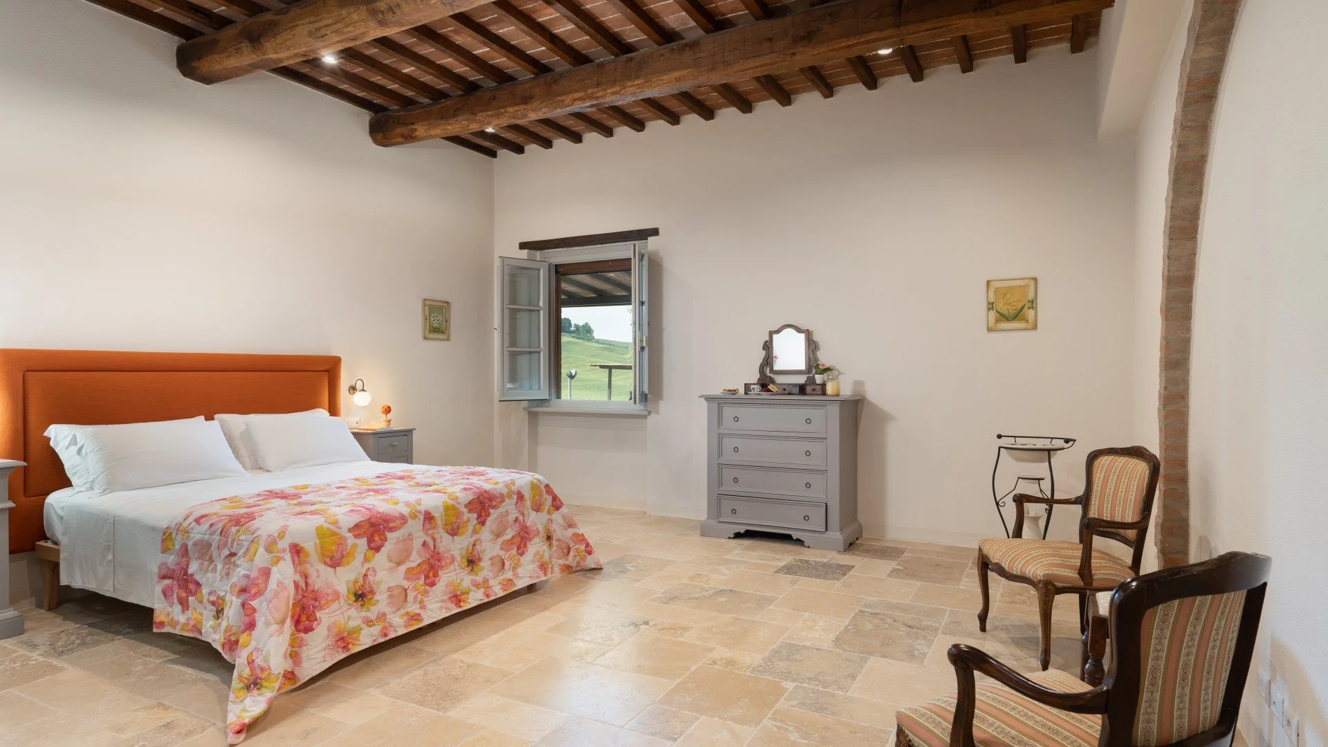 A bedroom with a bed featuring a floral bedspread, a gray headboard, and white pillows. There are two nightstands with bedside lamps, an open window with a view of green hills, and a gray dresser with a mirror. The room has tiled floors, exposed wood