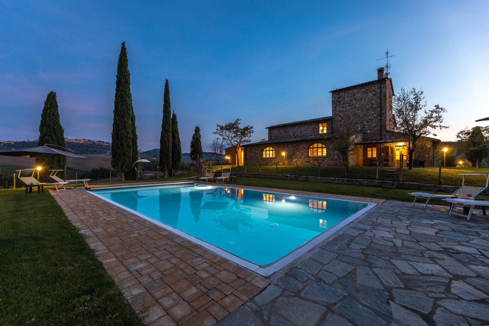 An outdoor swimming pool illuminated at night, with lounge chairs and umbrellas nearby, in front of a stone house with lit windows, surrounded by trees and tall cypress trees, with a hilly landscape in the background.