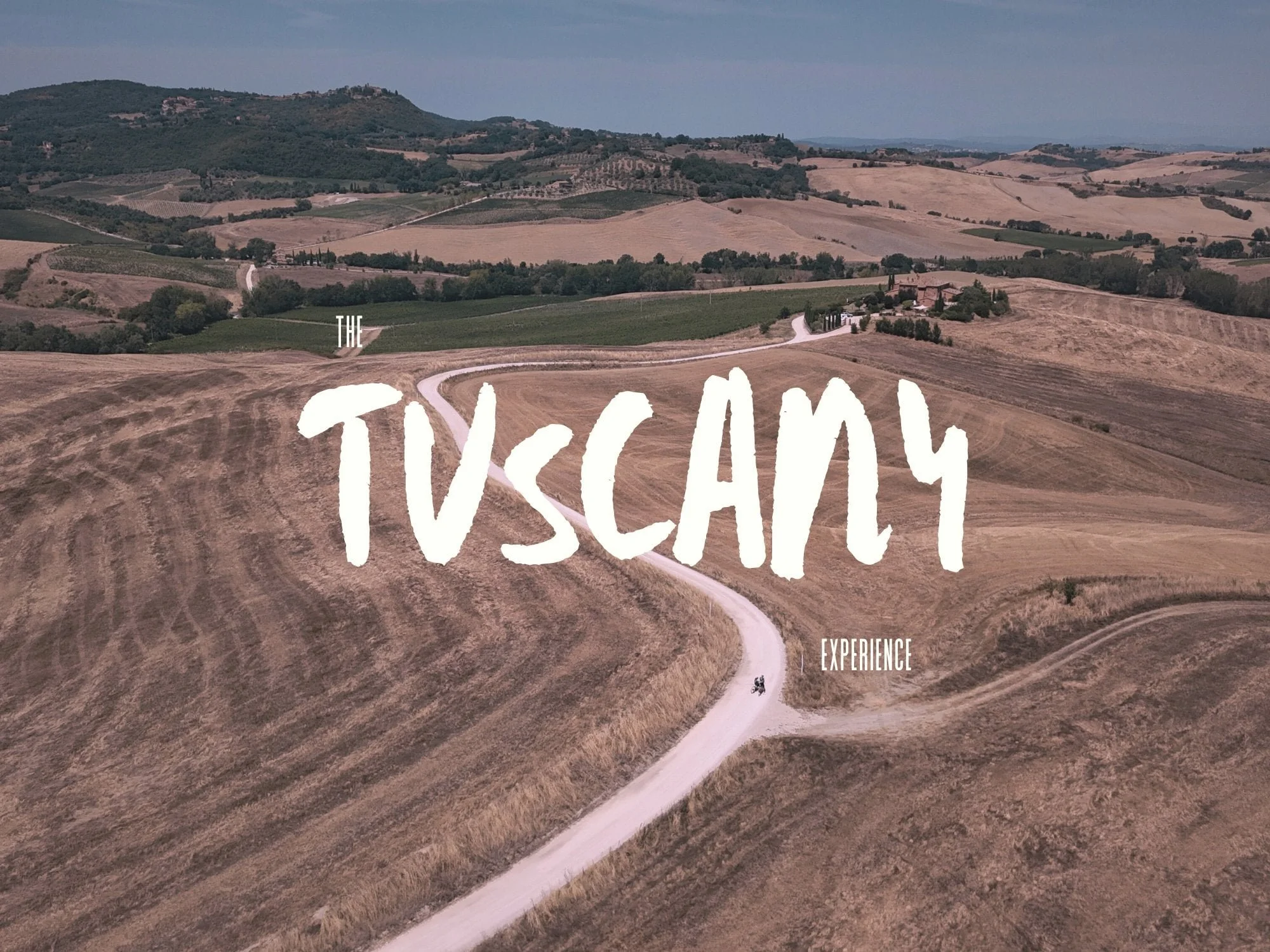 Aerial view of rolling hills and farmland with a winding dirt road, overlaid with the text 'The Tuscany Experience' in large white letters.
