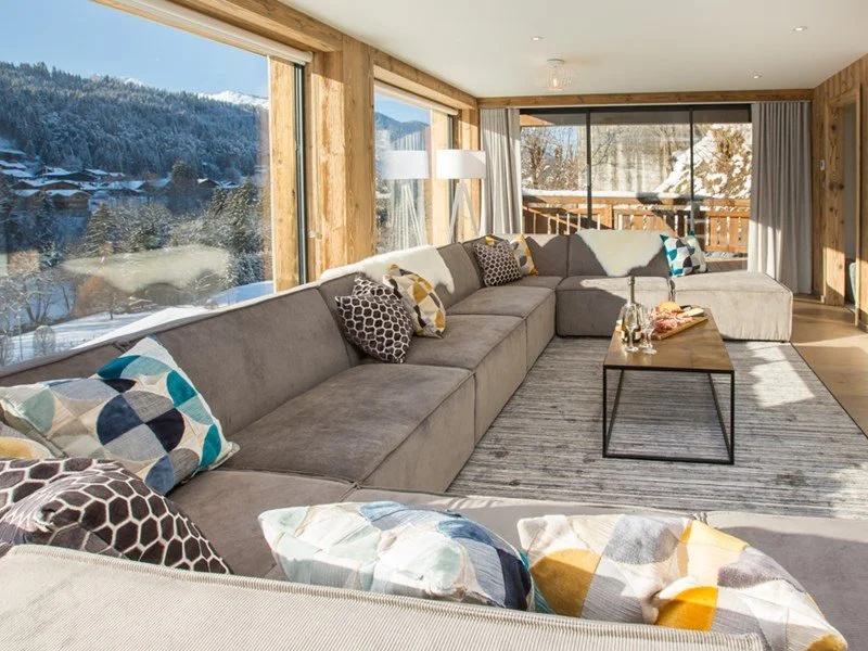 Cozy living room with a large gray sectional sofa adorned with patterned pillows, set in front of big windows showing a snowy mountain landscape, with a wooden coffee table holding drinks and snacks, and a sliding glass door leading to a balcony.