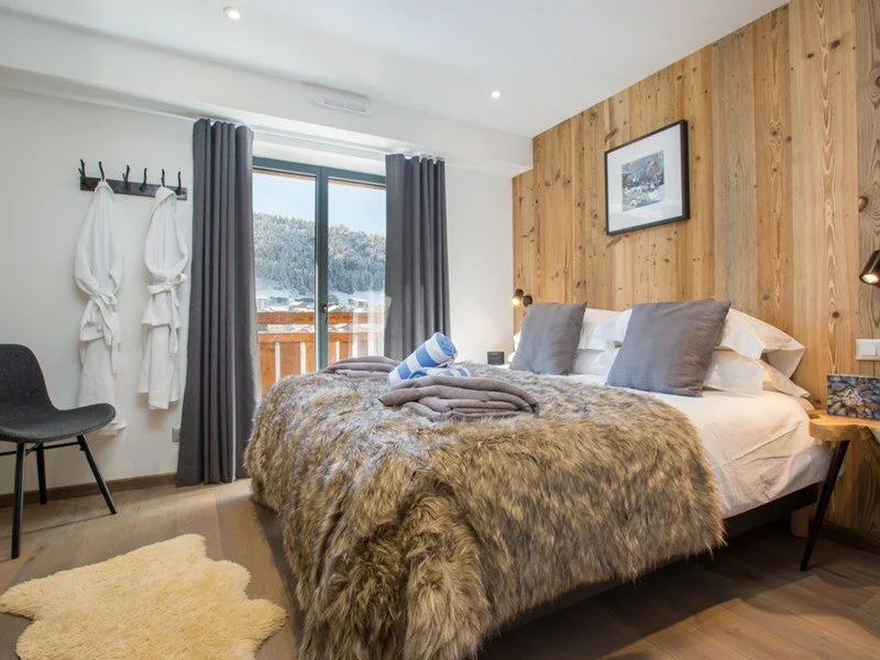 A cozy bedroom with a bed covered in a fur blanket, two gray pillows, and a striped towel. There's a wooden wall behind the bed, a framed picture, and a sliding glass door leading to a balcony with a scenic view of mountains and trees. To the left, j
