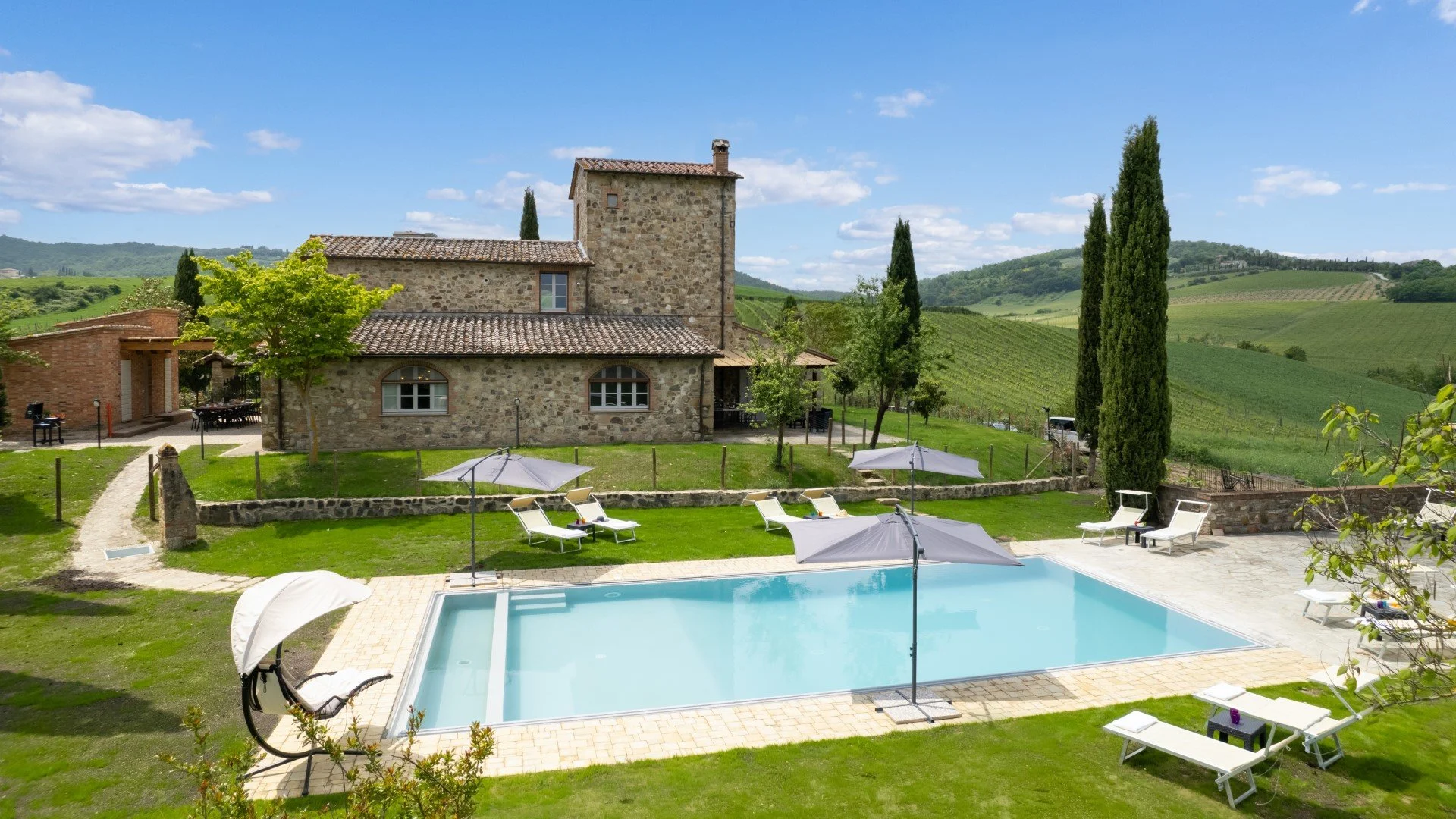 A scenic view of a stone house with a tiled roof, surrounded by greenery, cypress trees, and rolling vineyard hills in the background. In the foreground, there is a swimming pool with lounge chairs and umbrellas on a paved patio.