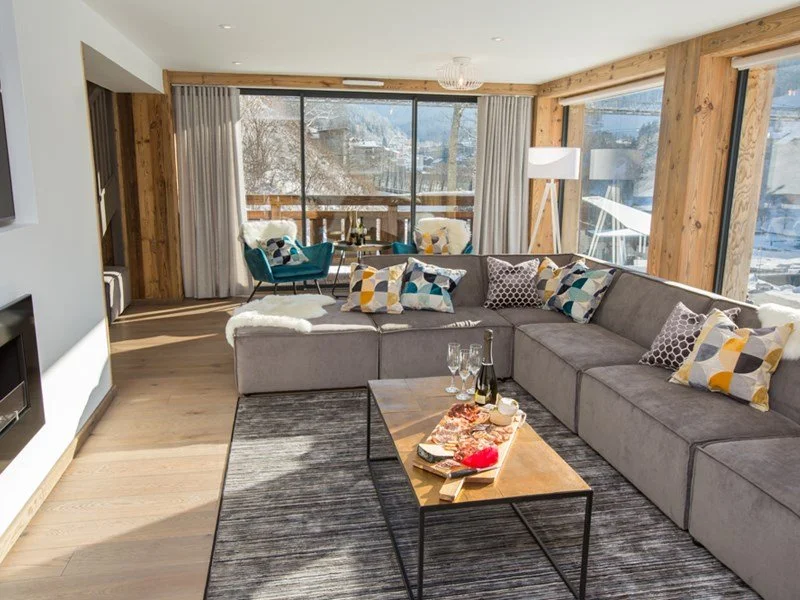 Modern living room with large window view of snowy mountains, gray sectional sofa with colorful patterned pillows, wooden coffee table with drinks and snacks, and a small seating area with turquoise chairs and a view of snowy landscape outside.