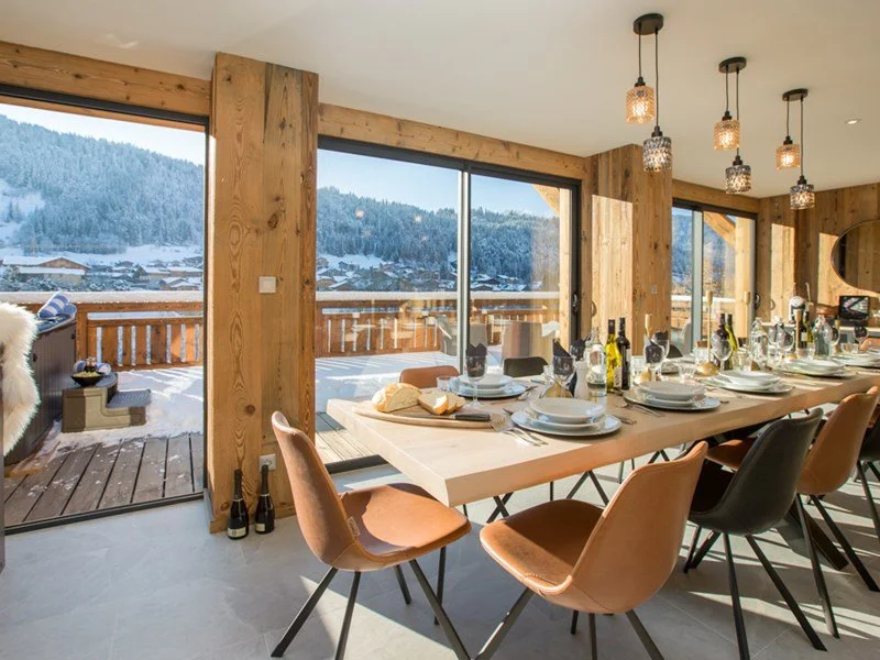 Modern dining room with a long table set for a meal, large windows showing a snowy mountain landscape outside, and pendant lights hanging from the ceiling.