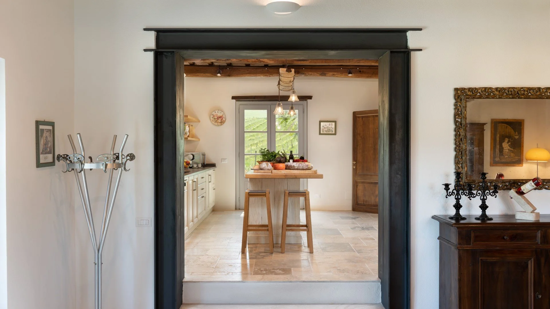 View of a cozy kitchen made of wood with a small center island and a window overlooking greenery, seen through a doorway in a home interior.