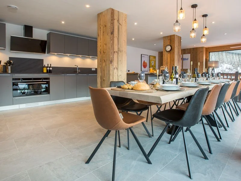 Modern dining room with a long table set with plates, glasses, bread, and bottles, surrounded by mixed brown and black chairs. The room features wood accents and contemporary hanging lights.
