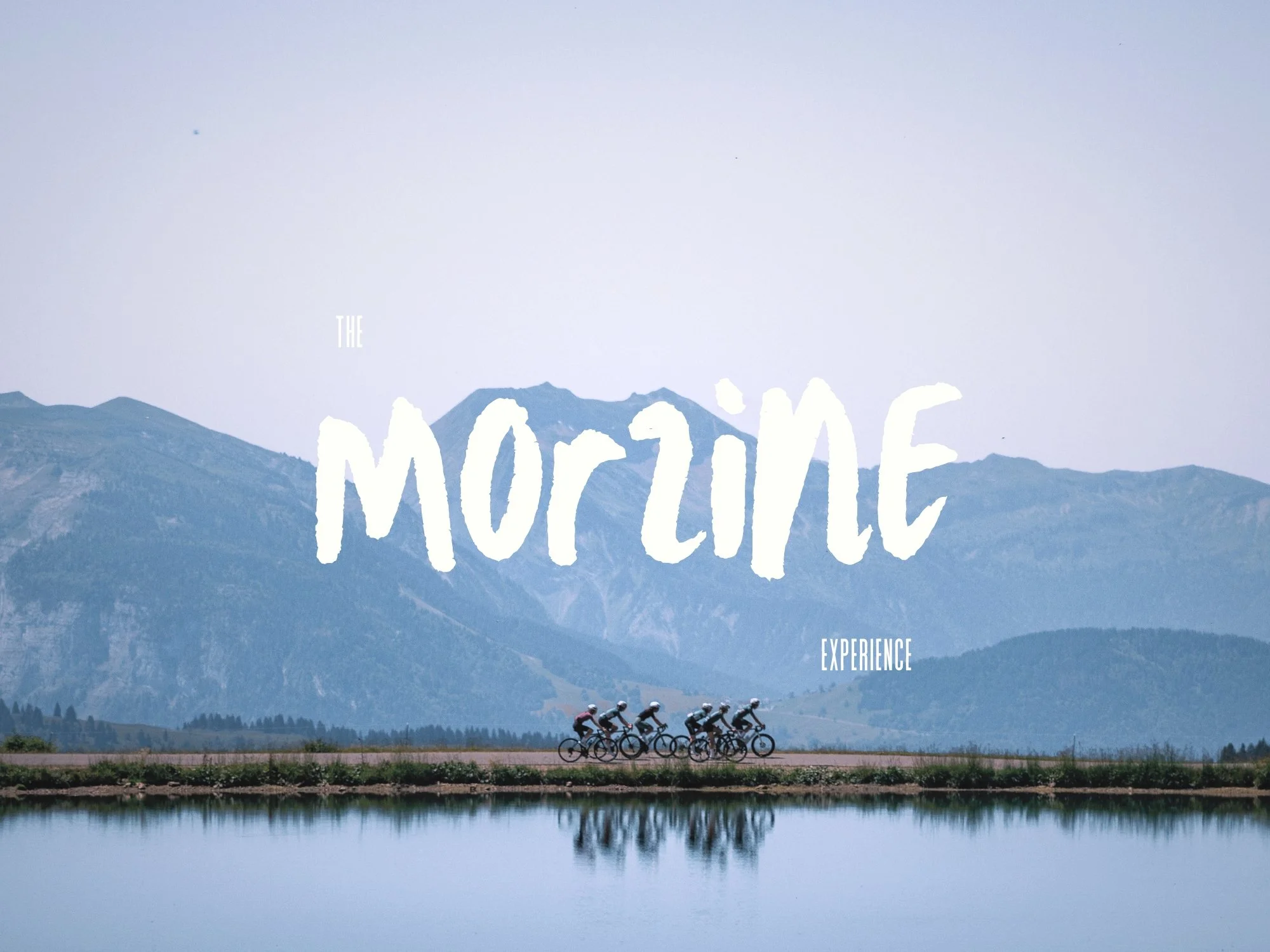A scenic mountain landscape with a group of cyclists riding on a road in front of a lake, with Spirit Mountain in the background. Overlaid text says 'The Mountain Experience'.
