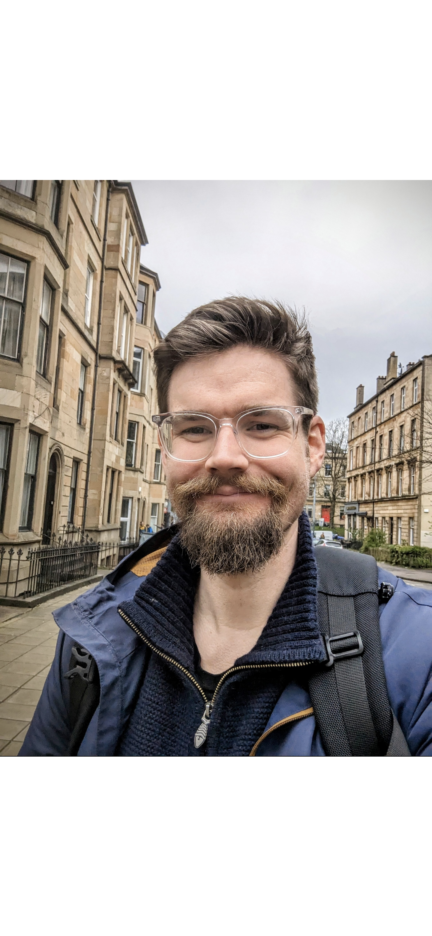 A smiling man with glasses and a beard taking a selfie on a city street with historic stone apartment buildings in the background.