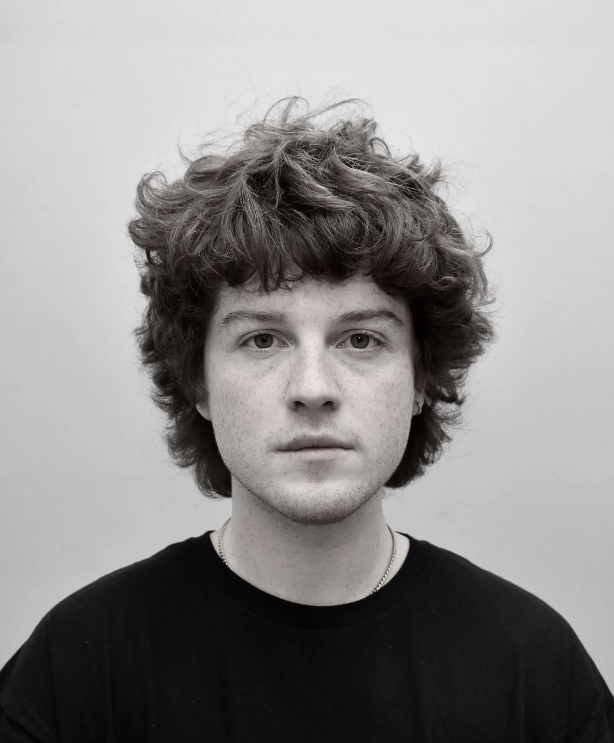 Close-up black-and-white photograph of a person with curly hair, visible eyes, nose, and mouth, wearing a dark shirt and a necklace, against a plain background.