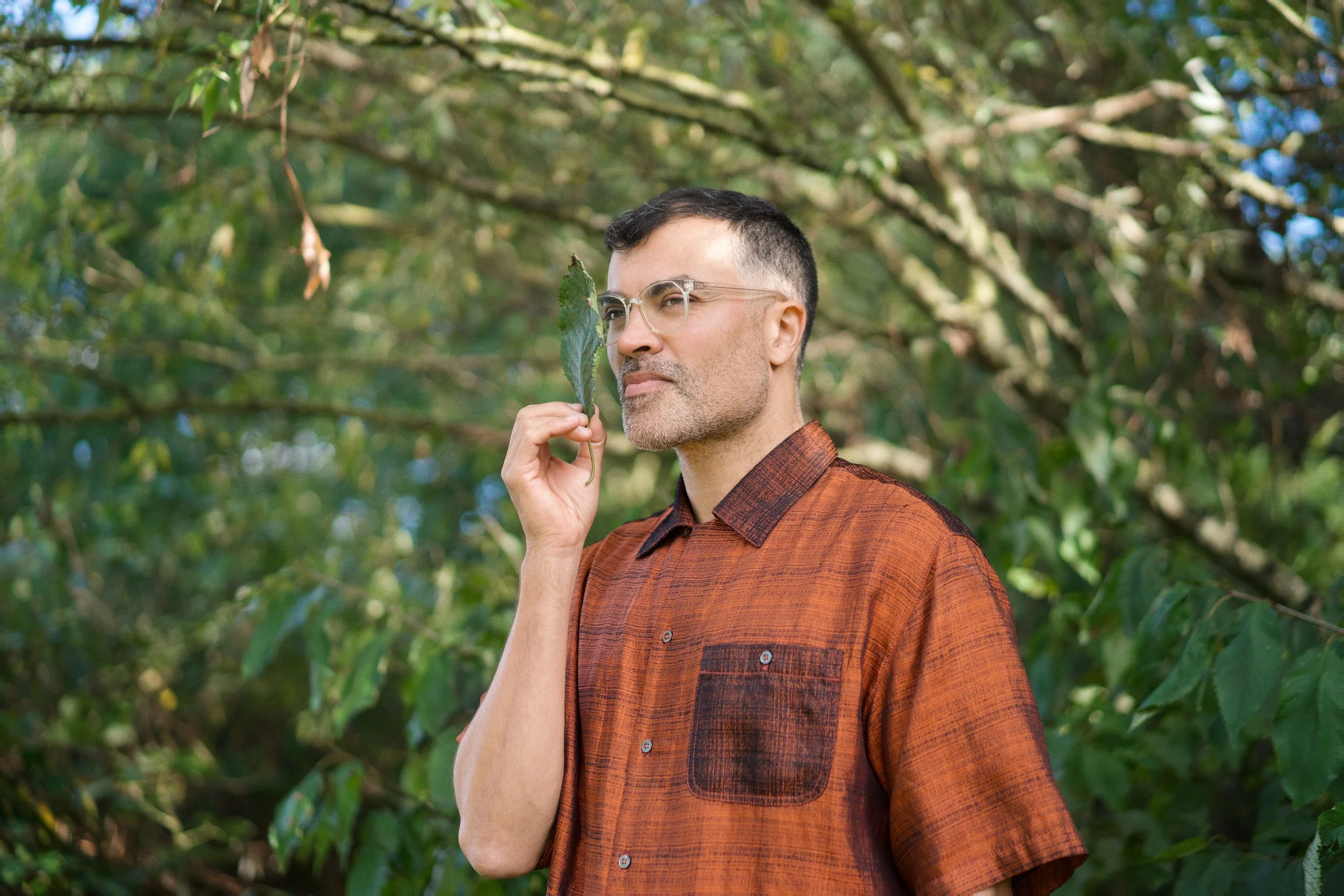 A man with glasses and short dark hair stands outdoors among green trees, holding a leaf close to his face.