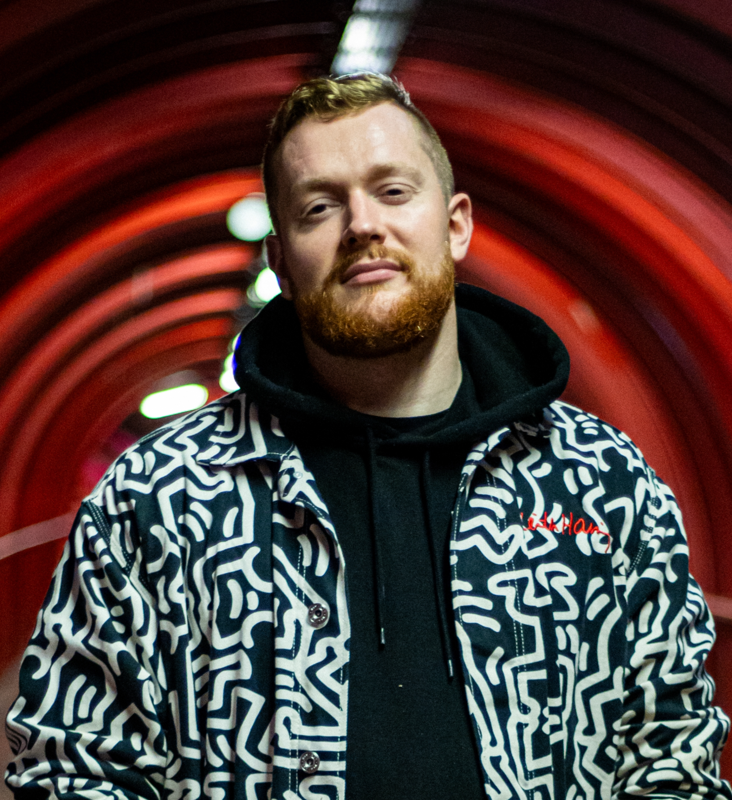 A young man with red hair, a red beard, and a red mustache is standing in front of a red and green glowing background with a tunnel-like effect. Tommy Dey, Creative Communities Producer