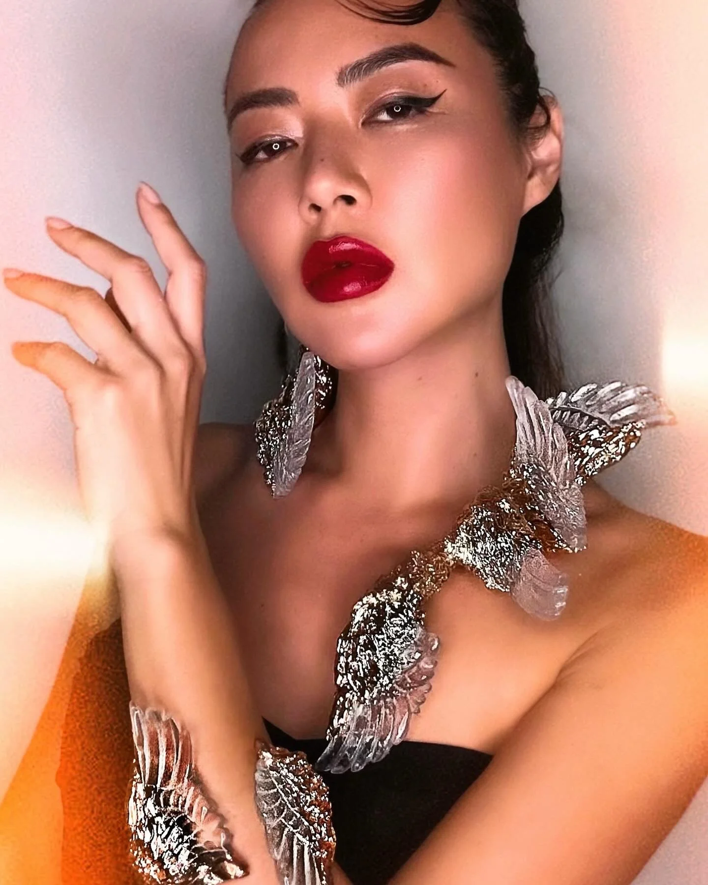 A woman with dark hair styled back, bold red lipstick, winged eyeliner, and eye shadow, wearing large silver butterfly-shaped earrings and a matching necklace. She has a confident expression and is touching her face with her left hand, against a plain background.