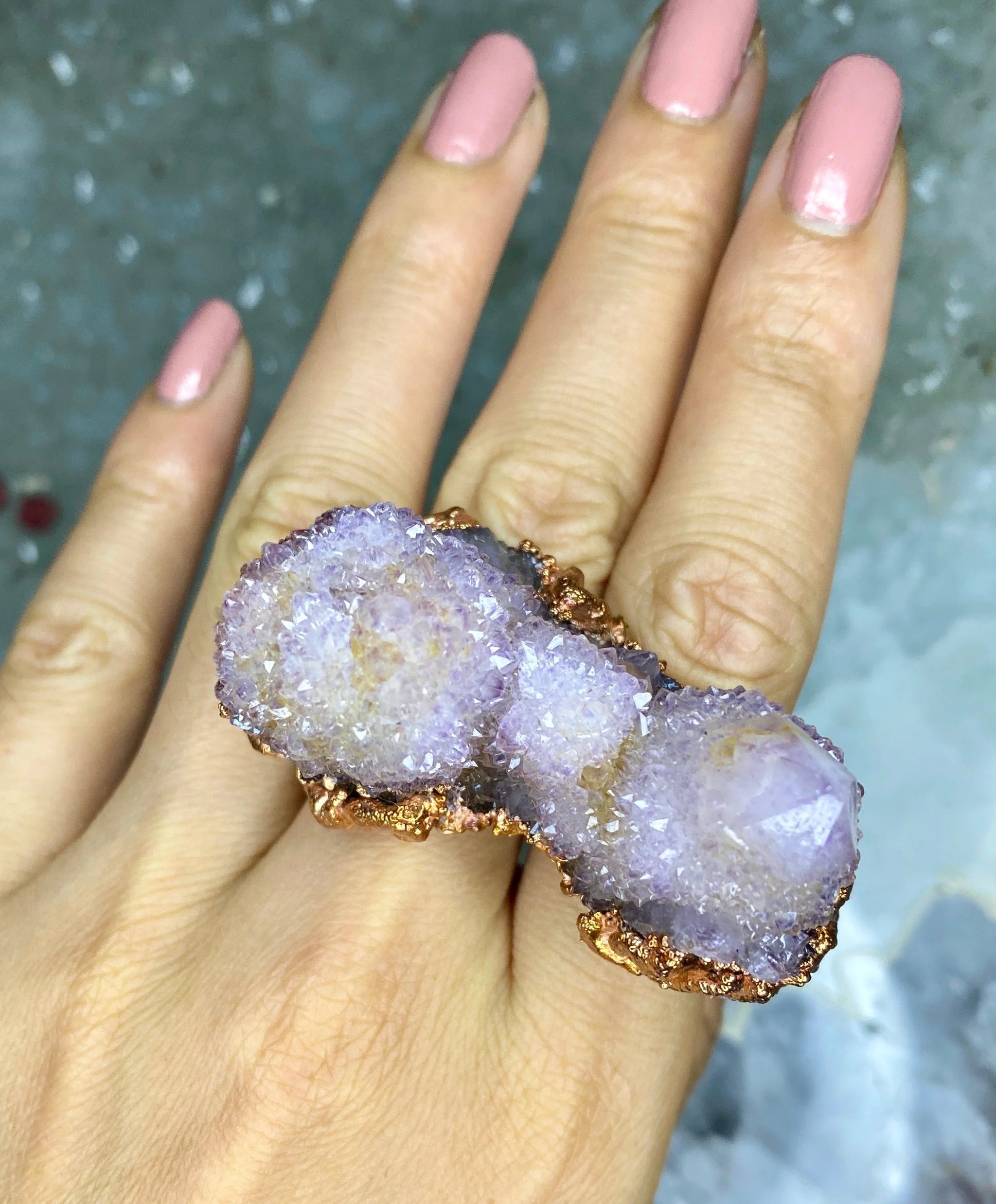 A large, sparkling ring with three purple geodes on a person's finger with pink painted nails.
