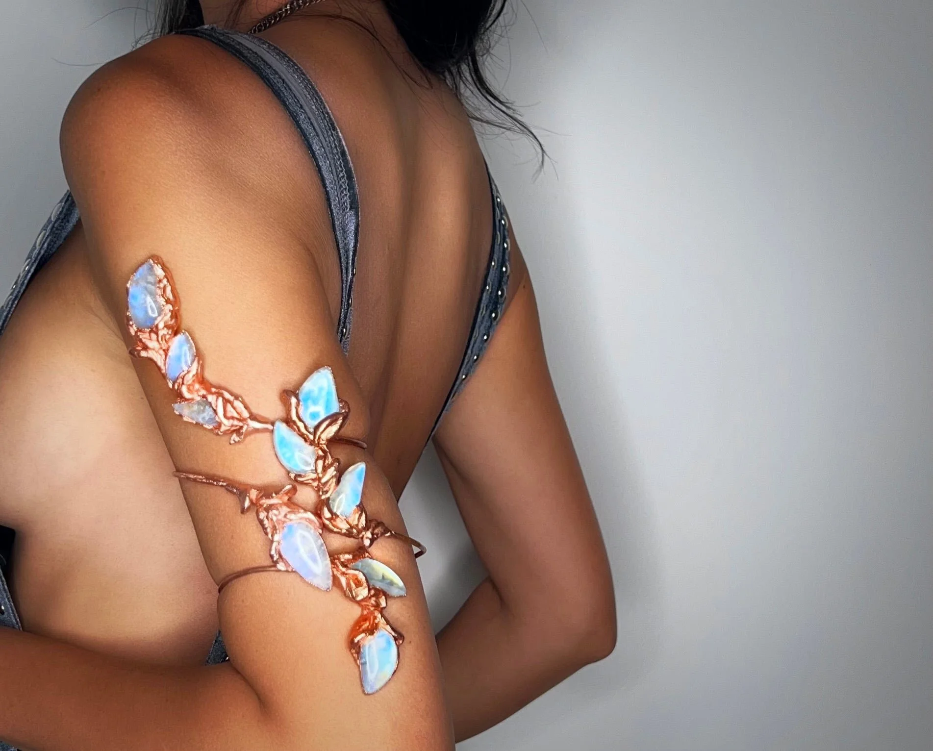 A woman with a detailed arm tattoo featuring a floral and bird design in blue and copper tones, wearing a dark gray tank top with metallic accents.