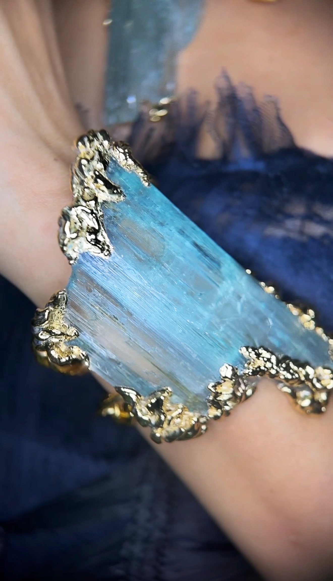 A person wearing a large bracelet with a large, rectangular, light blue gemstone with gold and black accents along the edges.
