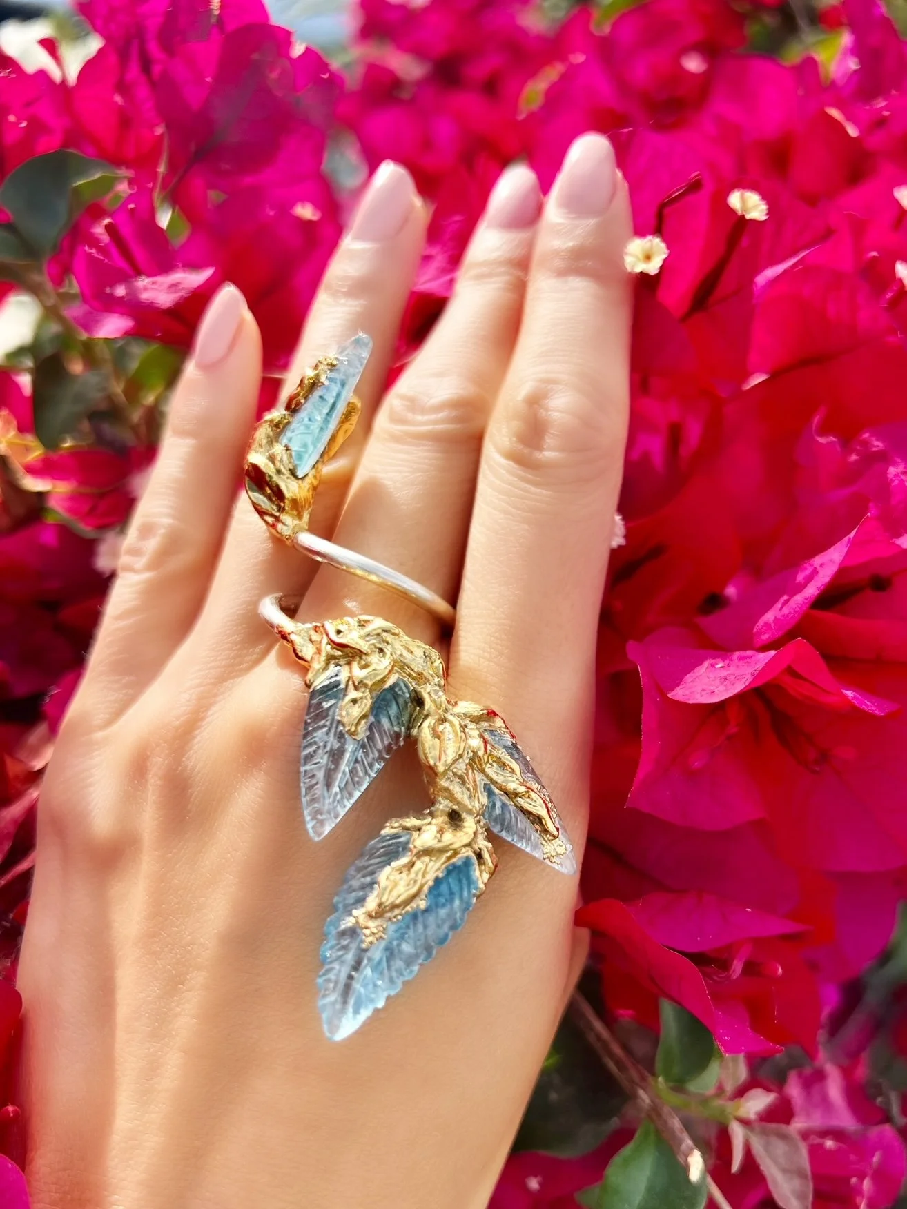 A hand with two rings, one on the middle finger and one on the ring finger, resting on bright pink flowers. The rings feature decorative details resembling birds with intricate wings, in gold and silver tones with blue accents.