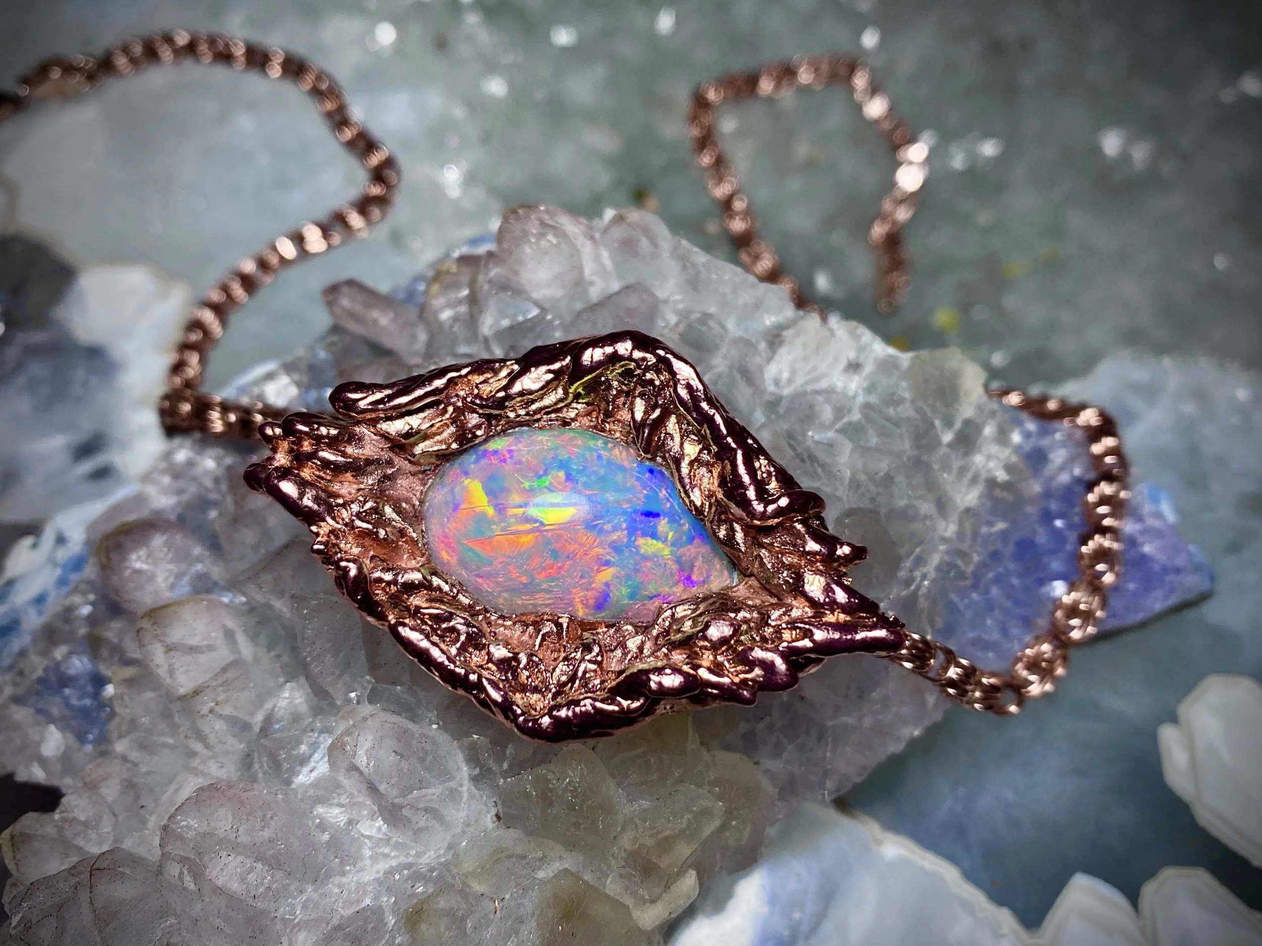 A close-up of a copper necklace with an oval opal gemstone, resting on a bed of white and blue crystal stones.