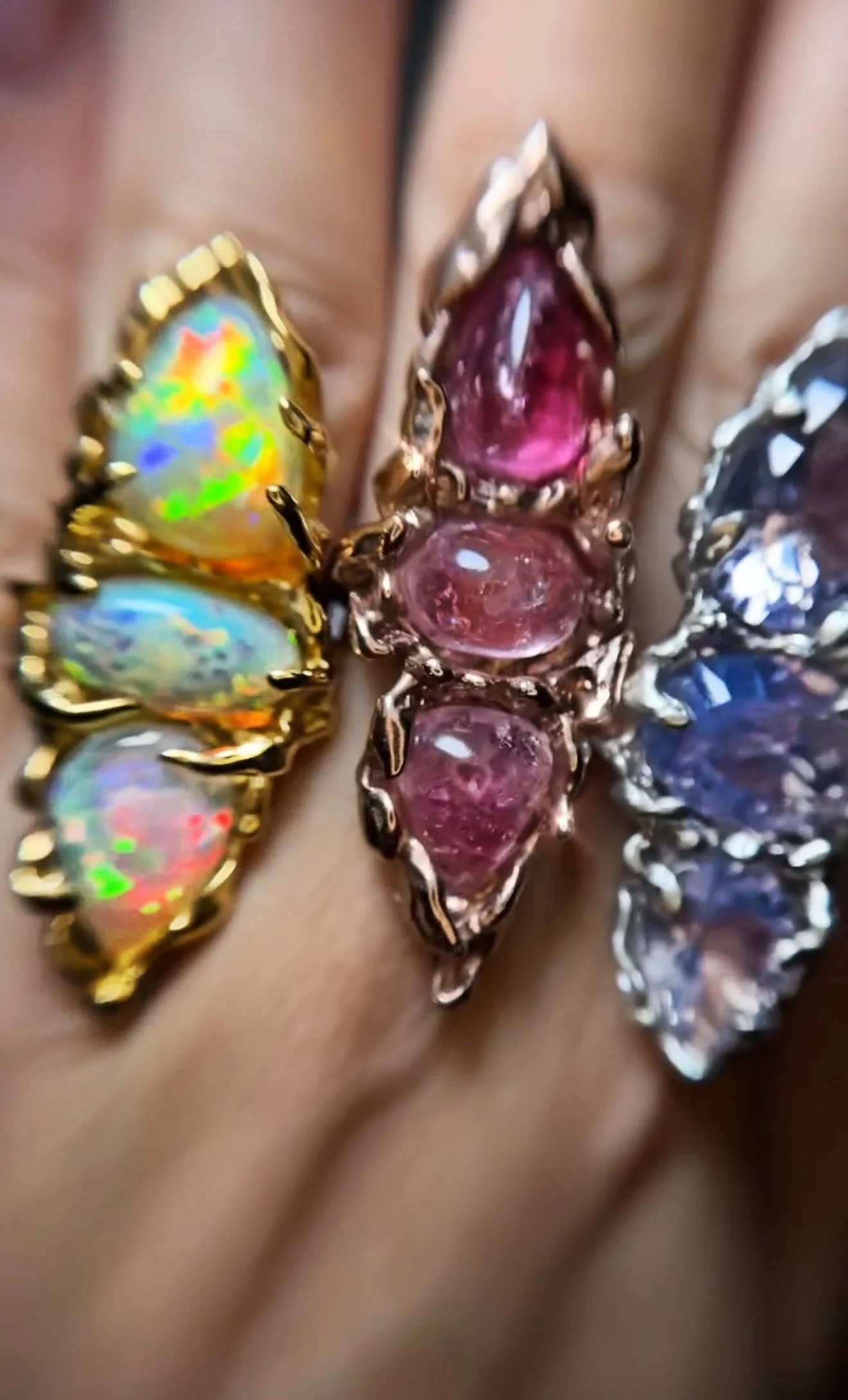 Close-up of three colorful gemstone rings on fingers, featuring a butterfly-shaped gemstone with iridescent green, blue, yellow, and red hues, a pink gemstone ring, and a blue gemstone ring.