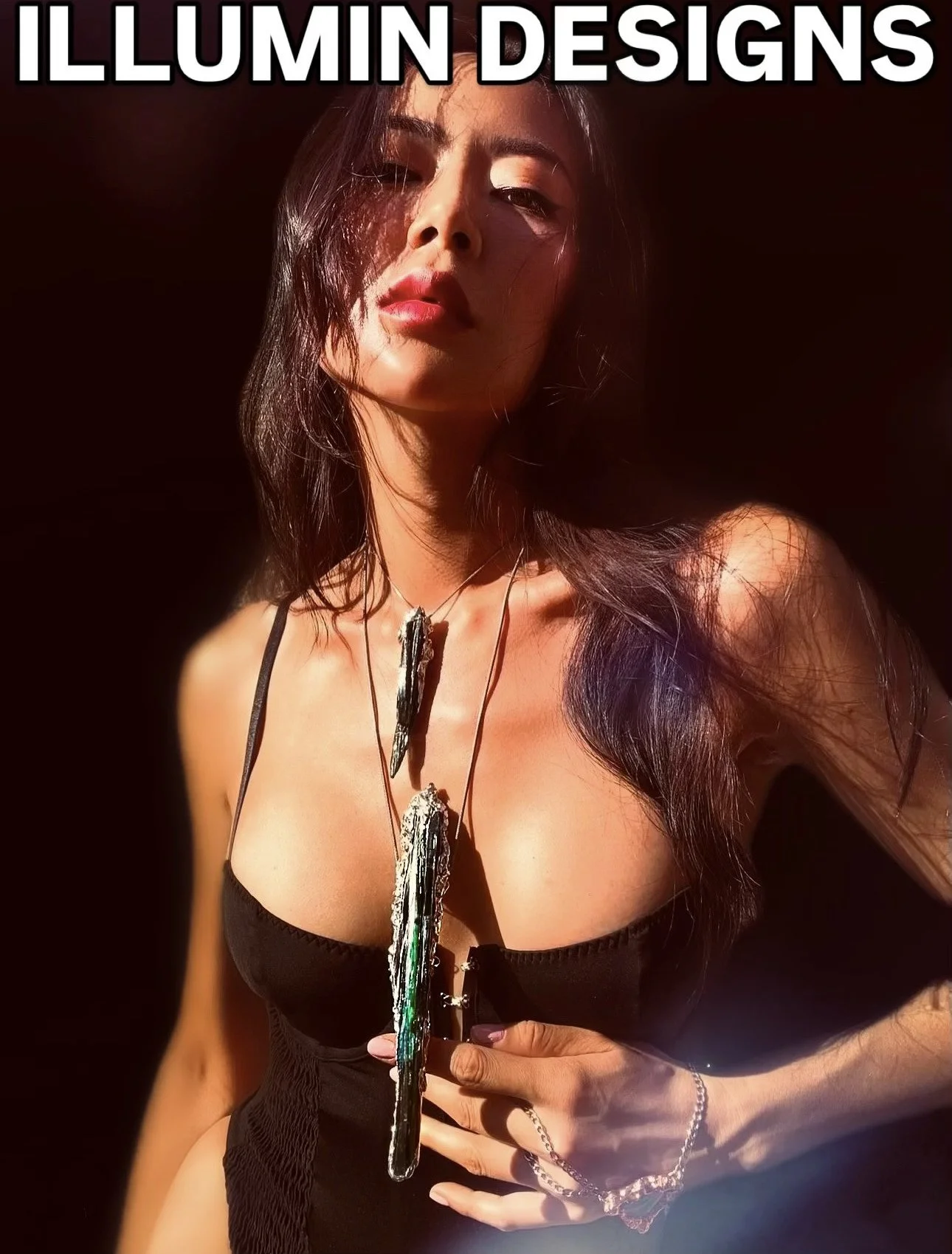 A woman with long dark hair posing against a dark background, wearing a black top, jewelry, and holding an object resembling a piece of wood or bark. She has a confident expression and is illuminated by light.