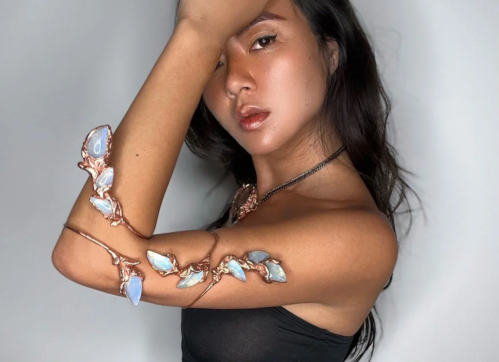 A woman with long dark hair, wearing metallic jewelry with blue and white stones, with one arm raised and hand on her forehead, against a plain background.