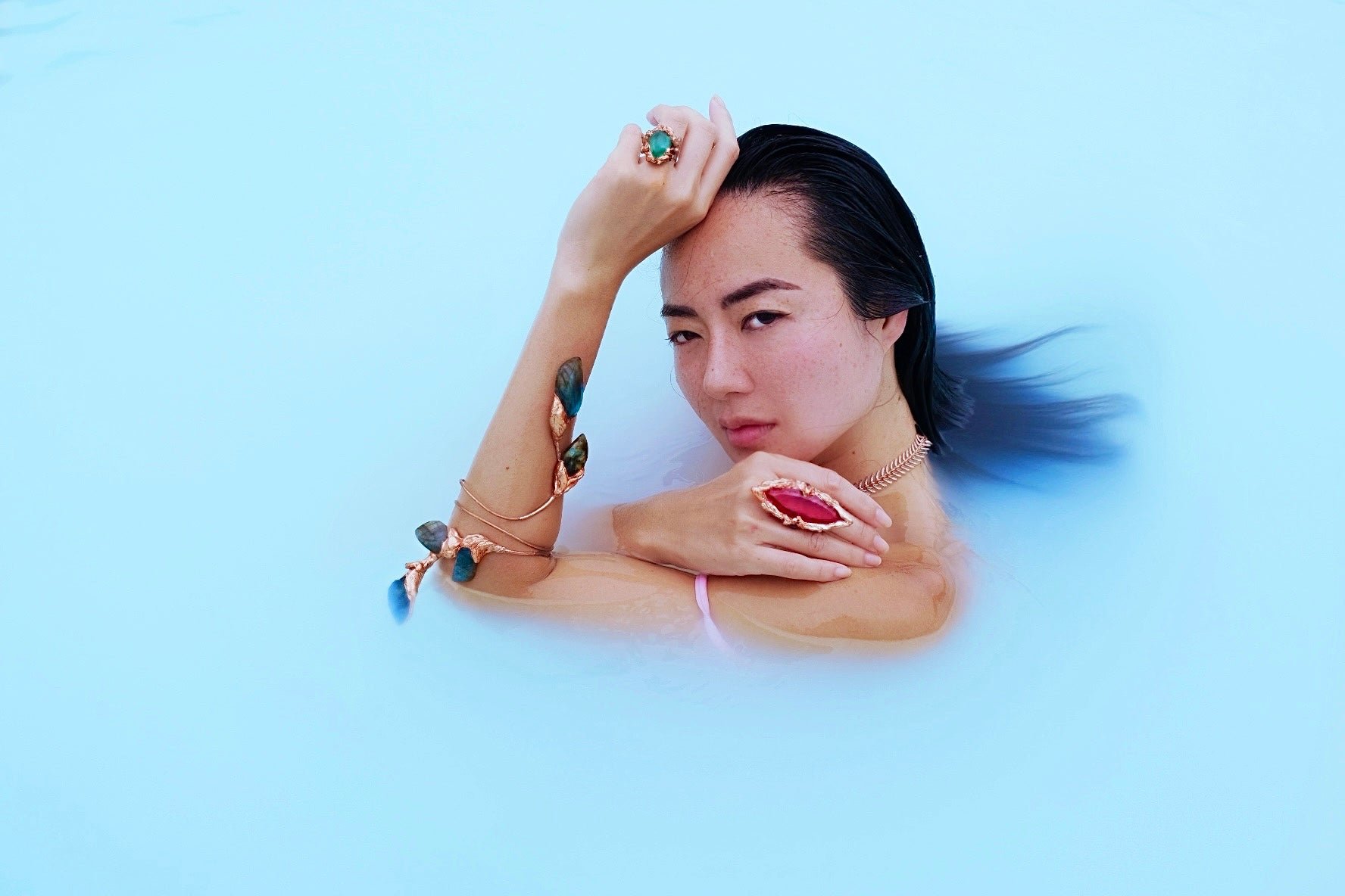 A woman with dark hair partially submerged in a light blue liquid, wearing multiple jewelry pieces including rings, necklaces, and bracelets, with her hand resting on her shoulder and her hair flowing in the water.