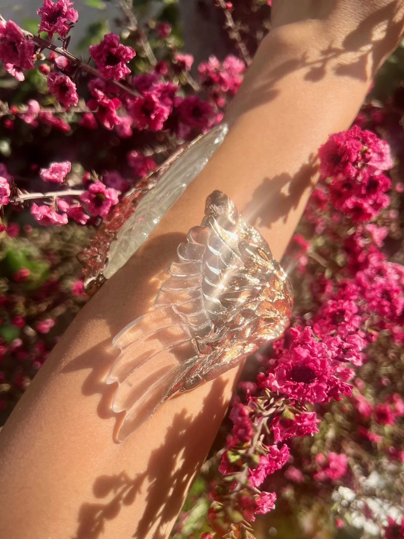 A person's arm with a silver feather-shaped bracelet, surrounded by pink flowers and sunlight.