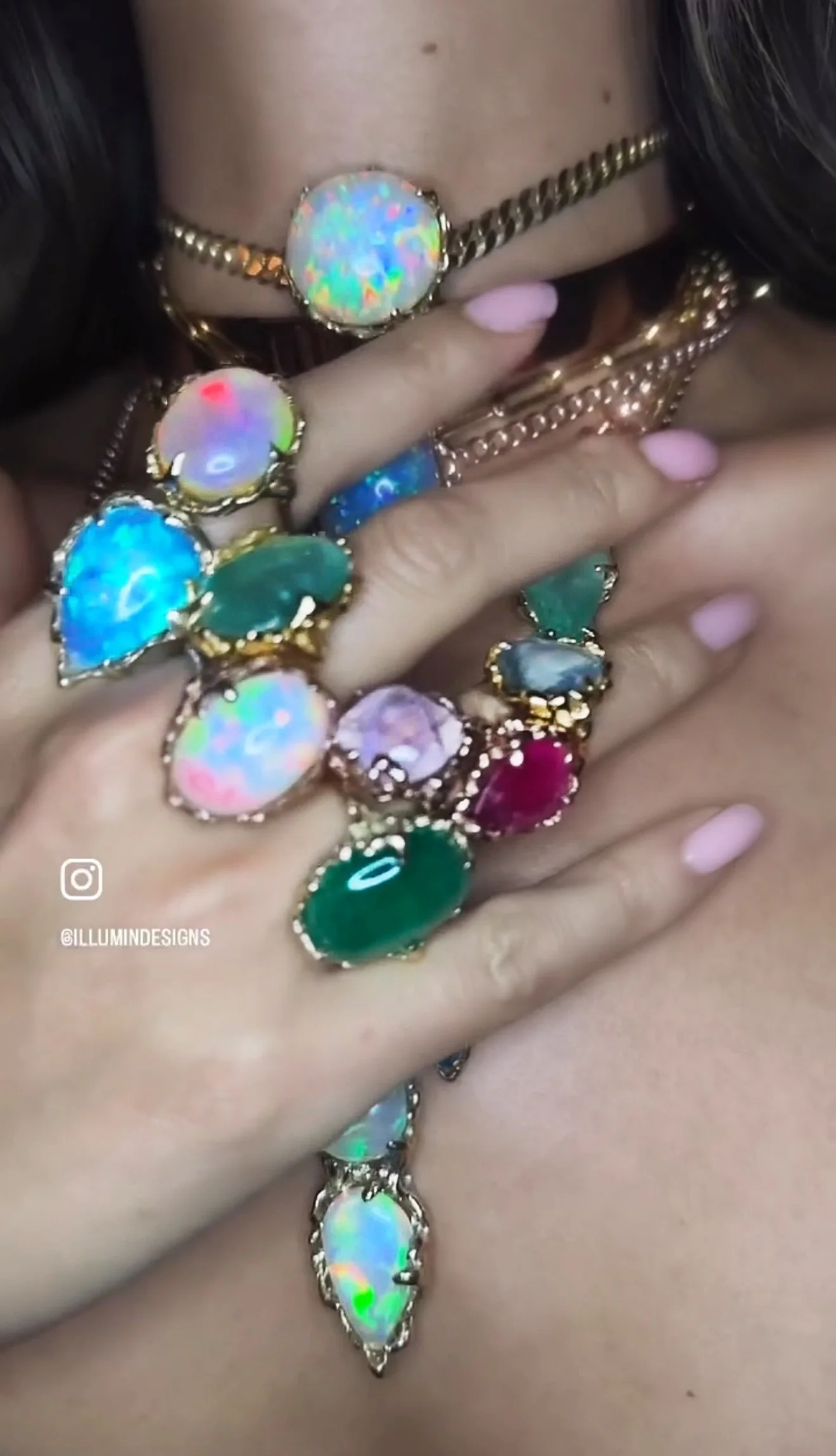 Person wearing multiple rings with large colorful opal gemstones, holding their fingers up near their face and chest. Some rings are gold, others are silver, with pink nail polish on their fingers.