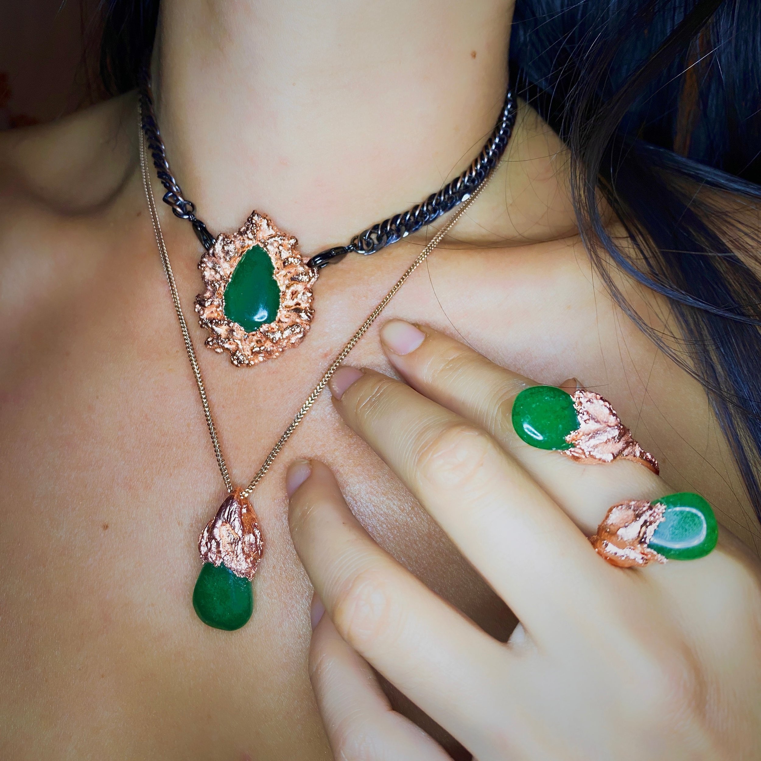 Close-up of a woman wearing green jade jewelry and a black and copper necklace, with matching rings and pendants, holding her hand near her chest.
