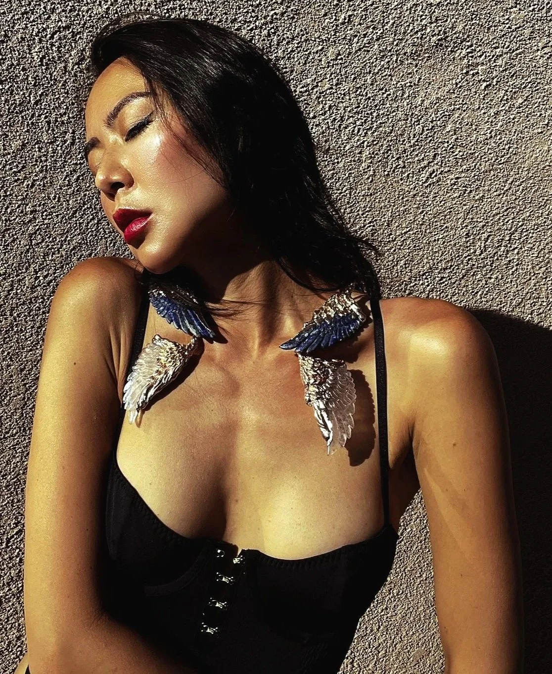 A woman with dark hair wearing a black top with a button-up front and large angel wing accessories on her shoulders, striking a pose against a textured wall with her eyes closed and head tilted.