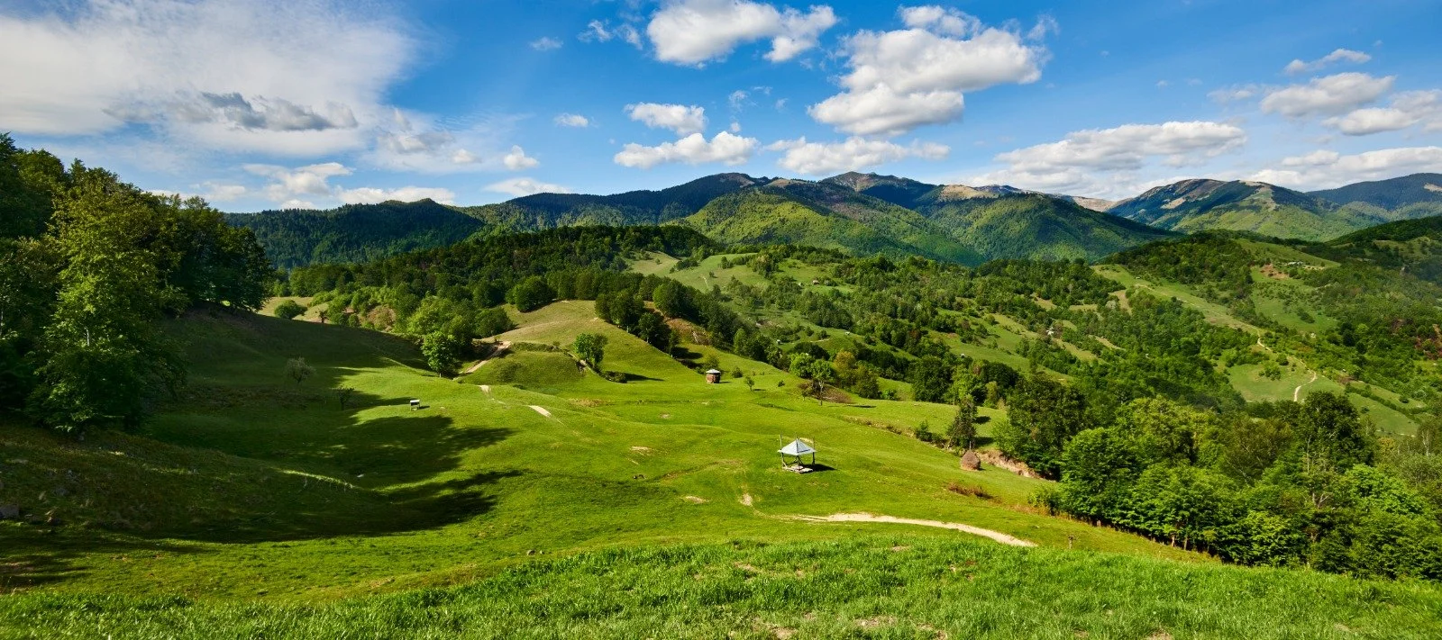 Scenic landscape with green rolling hills, small huts, and forested areas under a blue sky and scattered clouds, with distant mountains in the background.