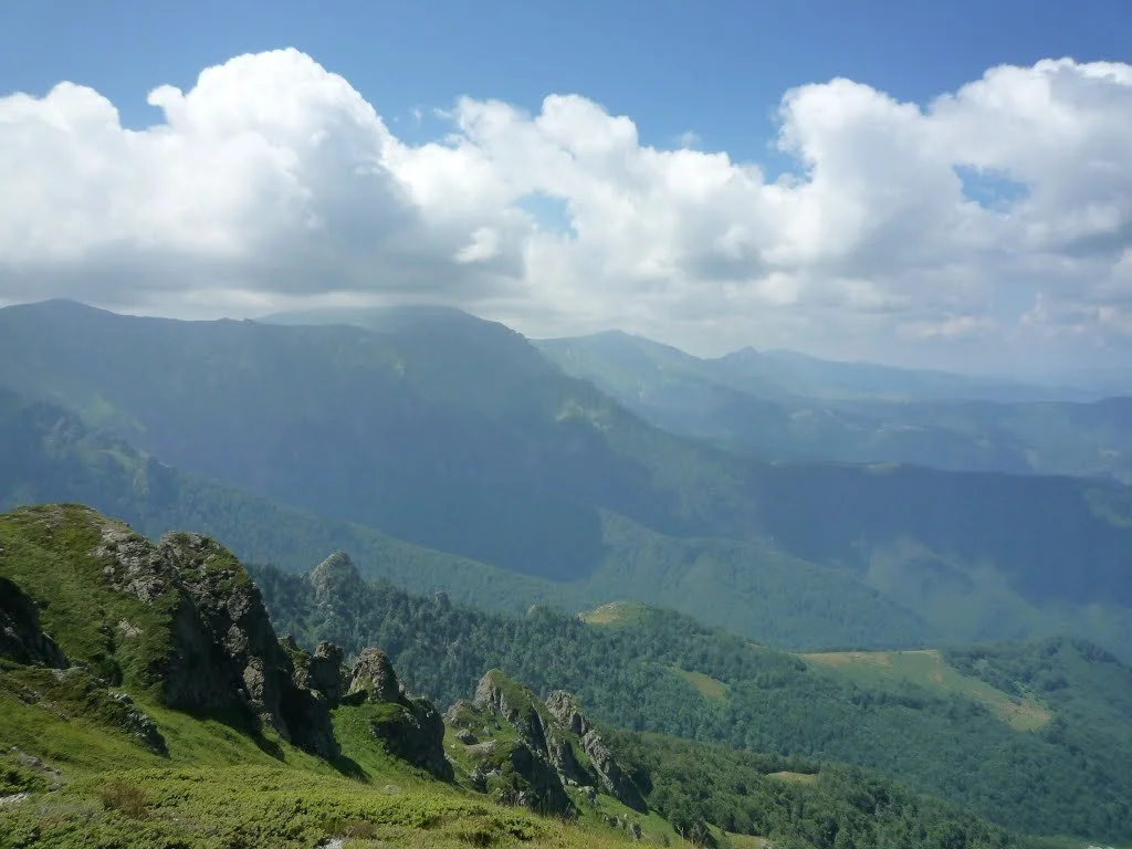 Mountain landscape with rocky peaks, green vegetation, and a cloudy sky.