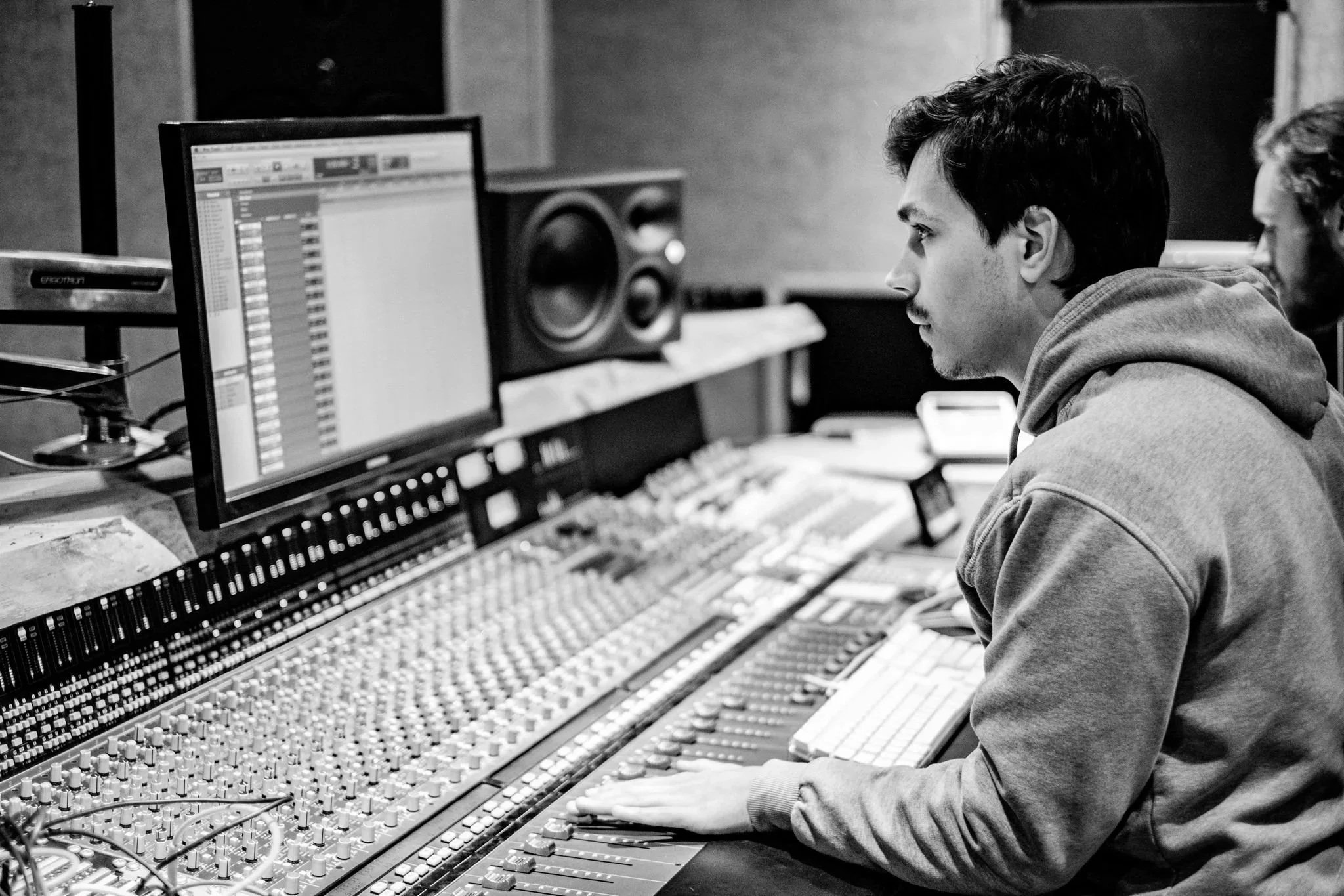A young man working at a music mixing console in a recording studio, focused on a computer screen and wearing a hoodie.