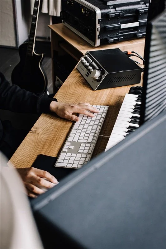 A person working at a wooden desk with a computer keyboard, a mixing console, and a keyboard instrument. Music equipment is visible, suggesting a music production or editing setup.