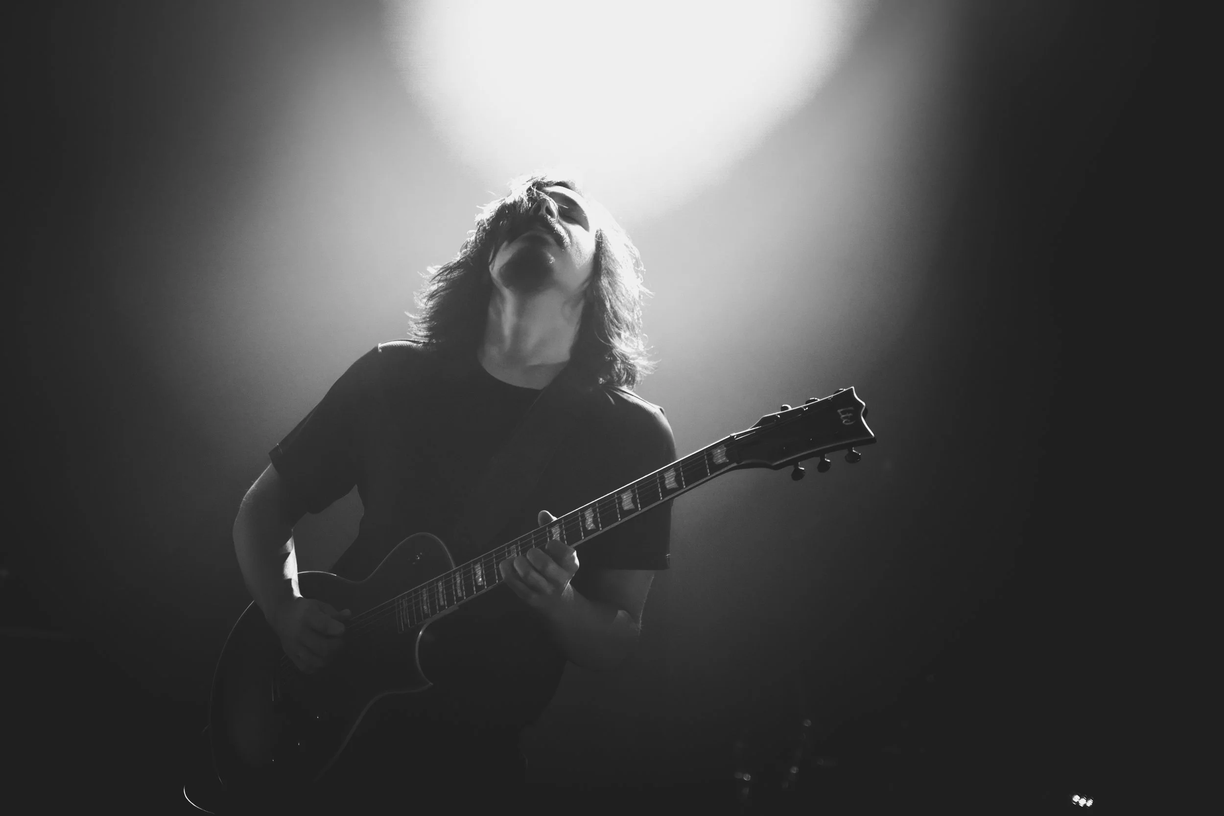 A person with long hair wearing sunglasses plays an electric guitar on stage, illuminated by bright overhead light in a black and white photo.