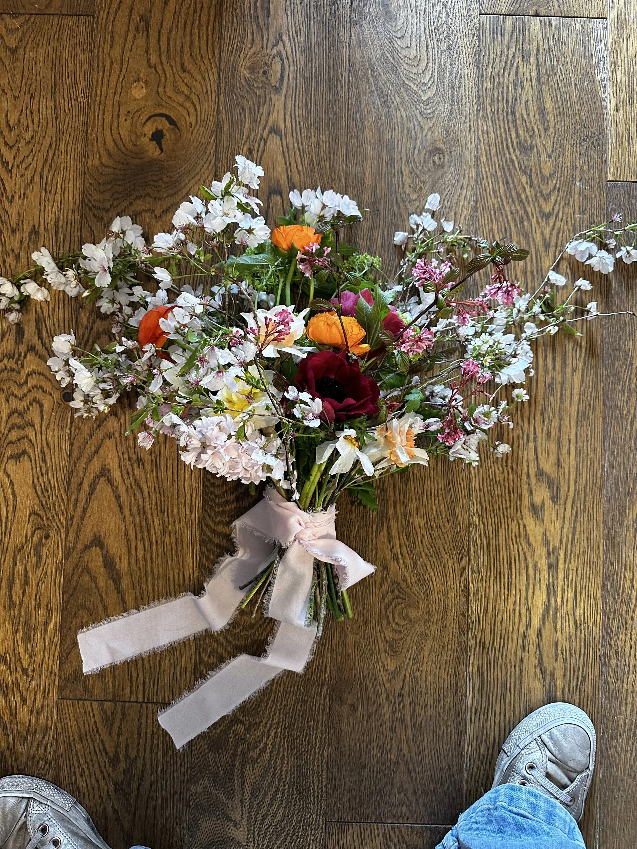 A colorful bouquet of flowers with white, orange, pink, red, and yellow blooms on a wooden floor, wrapped with a light pink ribbon.