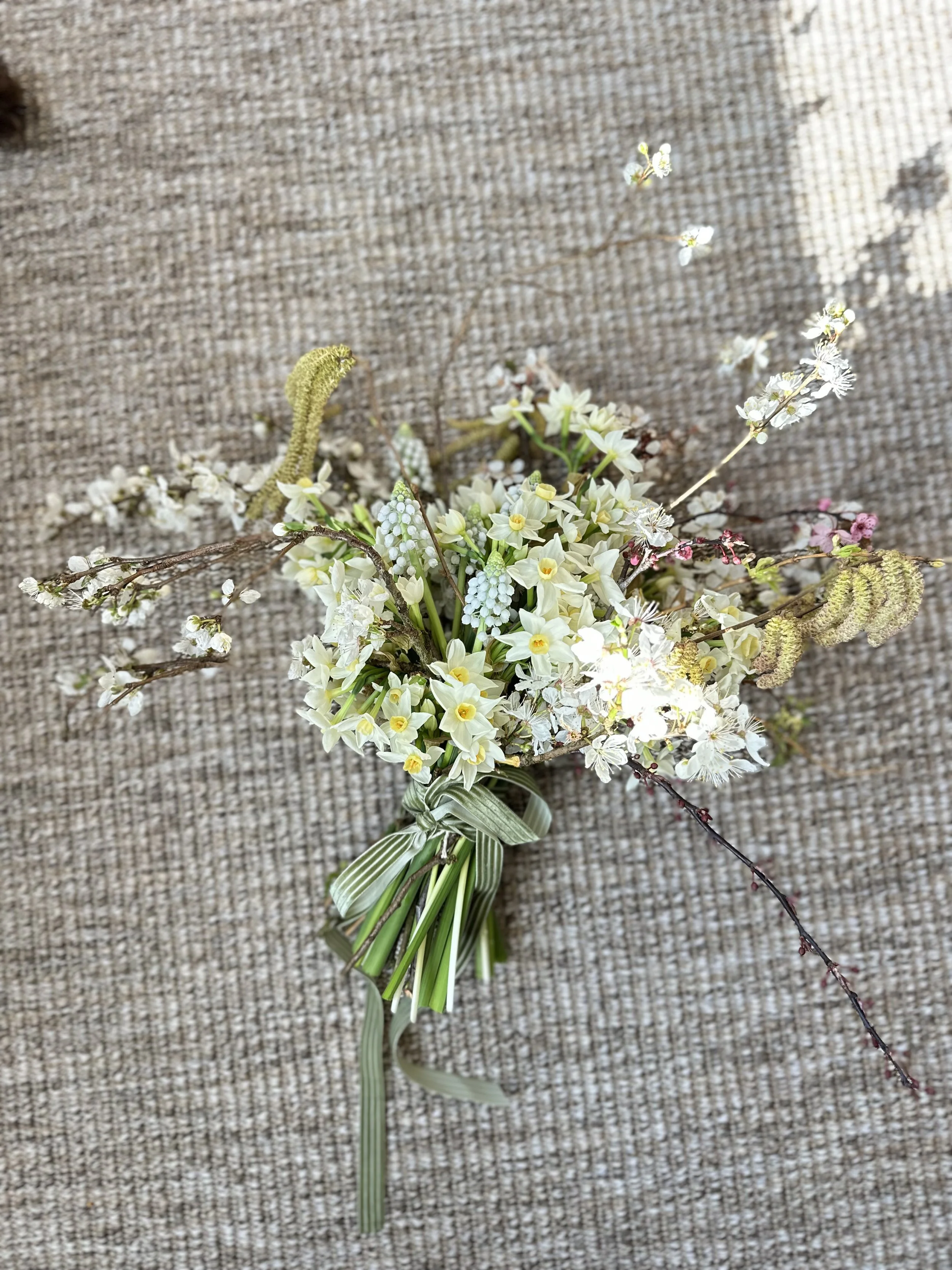 A bouquet of various small flowers including white and yellow ones, with some green and pink buds, tied together with a ribbon and placed on a textured beige surface.