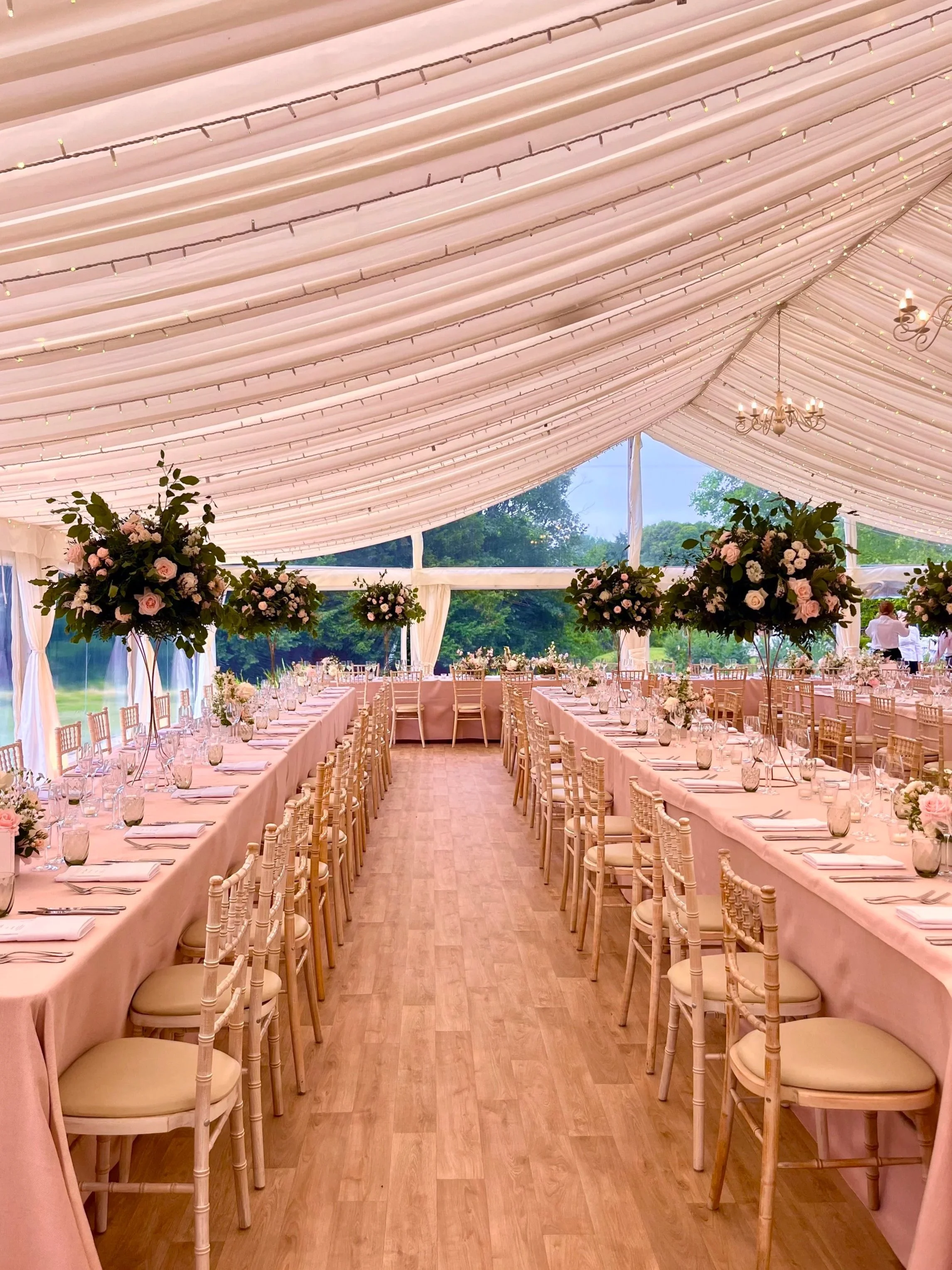 Elegant wedding reception tent with pink draped ceiling, hanging chandeliers, and long banquet tables decorated with pink and white floral centerpieces, set outdoors with green trees in the background.