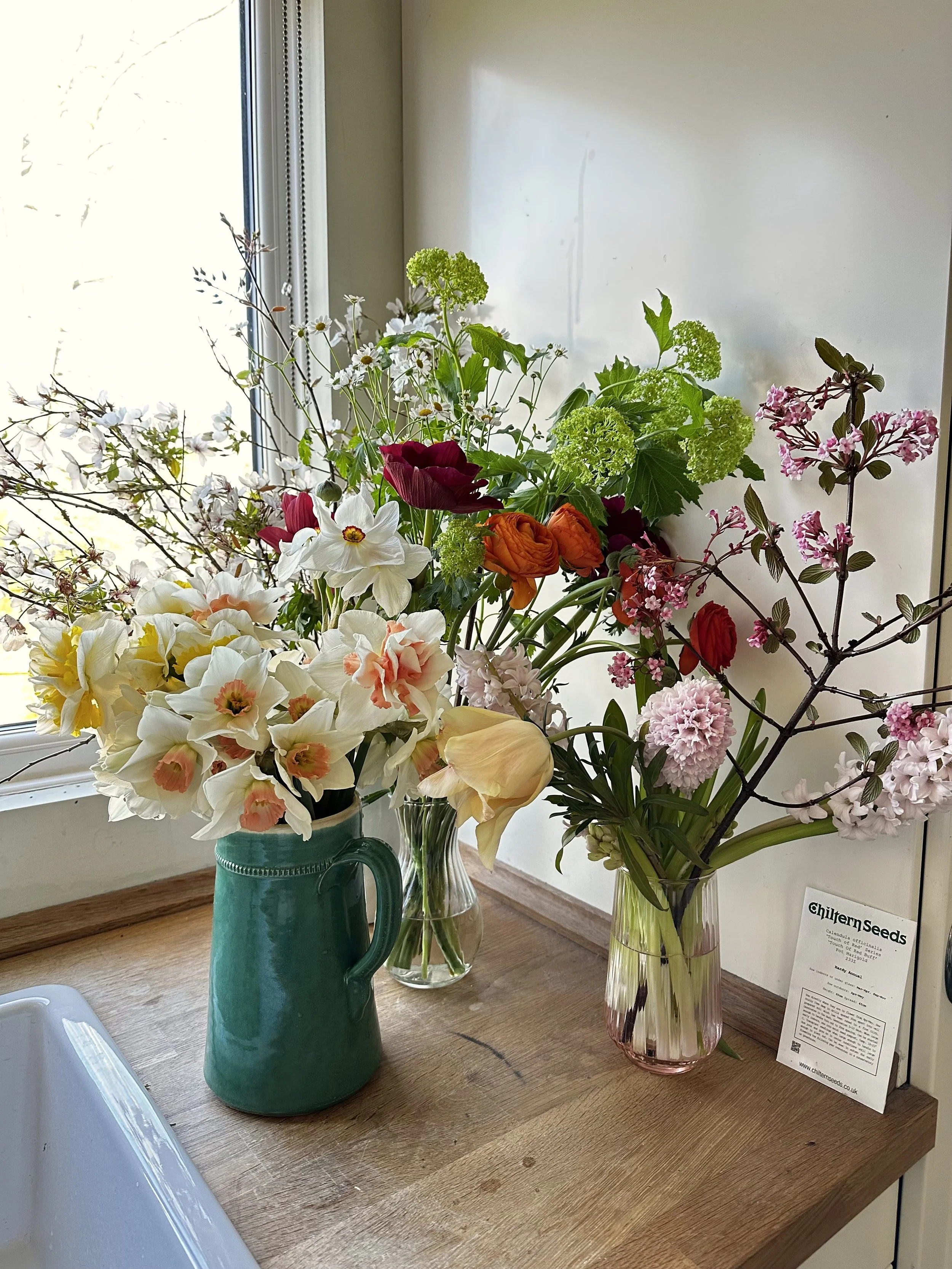 A collection of various flowers in vases on a wooden table near a window, including daffodils, lilies, roses, and other colorful blooms.