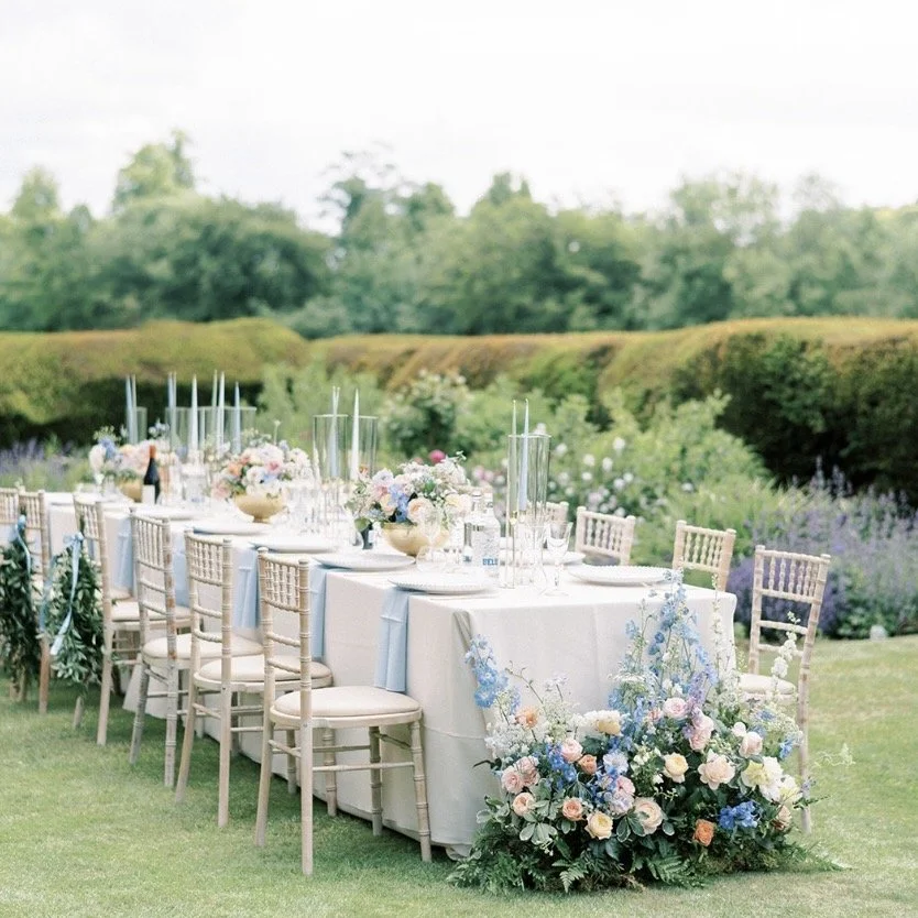 Outdoor wedding reception table setup with floral arrangements, candles, and elegant tableware on a lawn, surrounded by greenery.