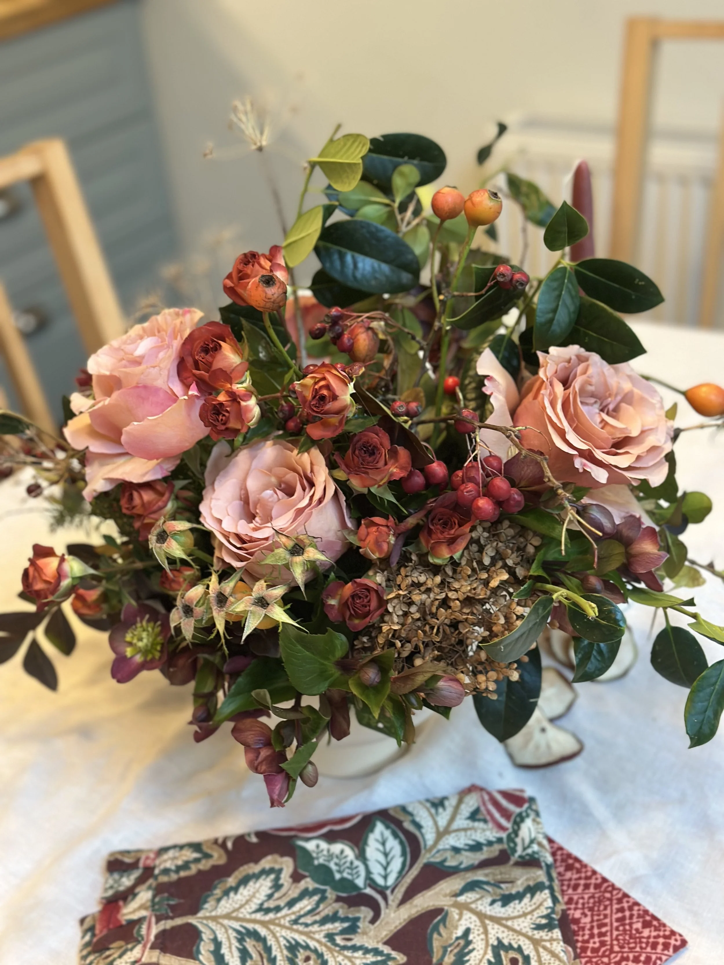 A floral arrangement with pink roses, small red berries, green leaves, and dried flowers, placed on a table with decorative cloths underneath.